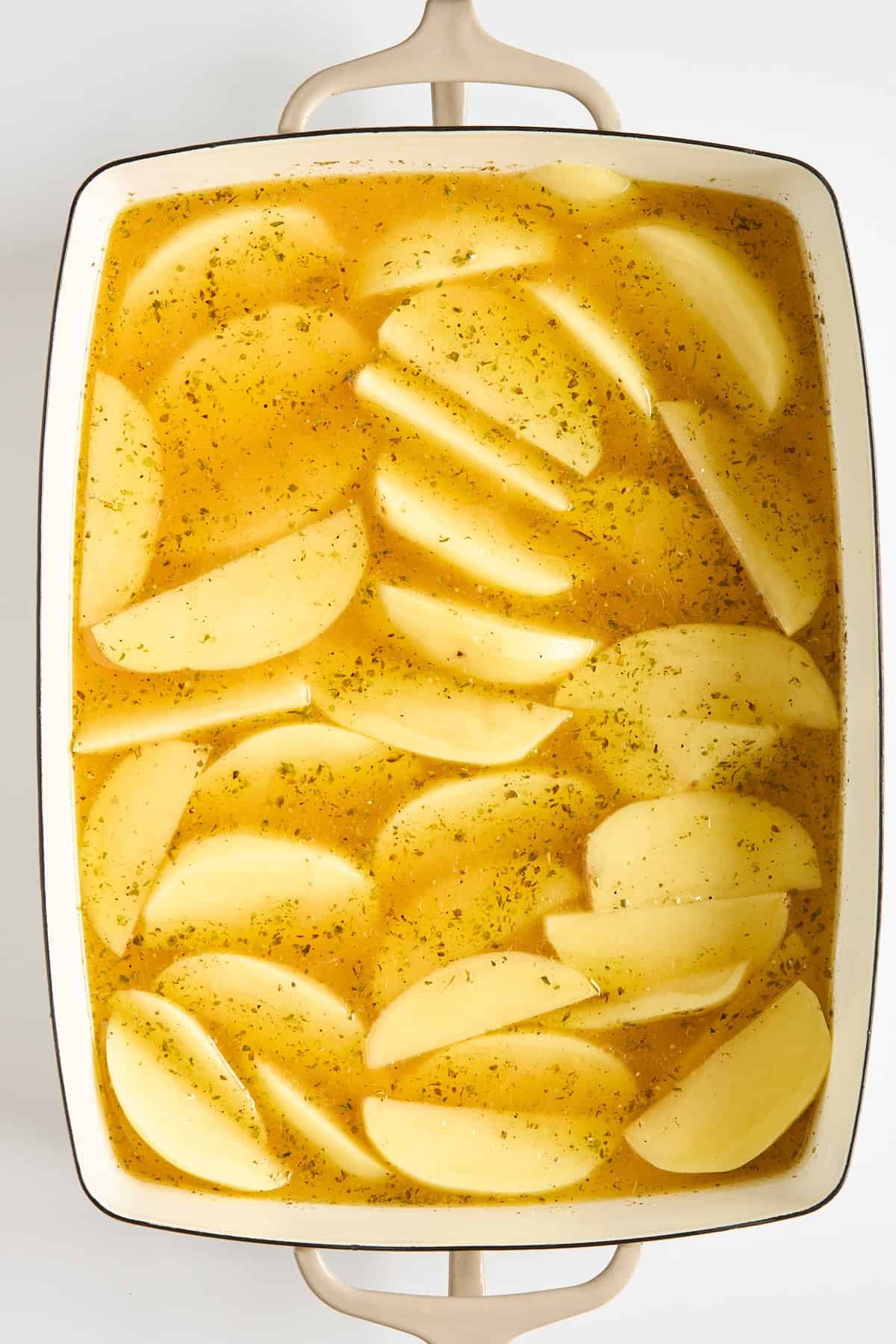 Sliced potatoes arranged in a white baking dish, covered in a yellow seasoned broth, ready to be cooked.