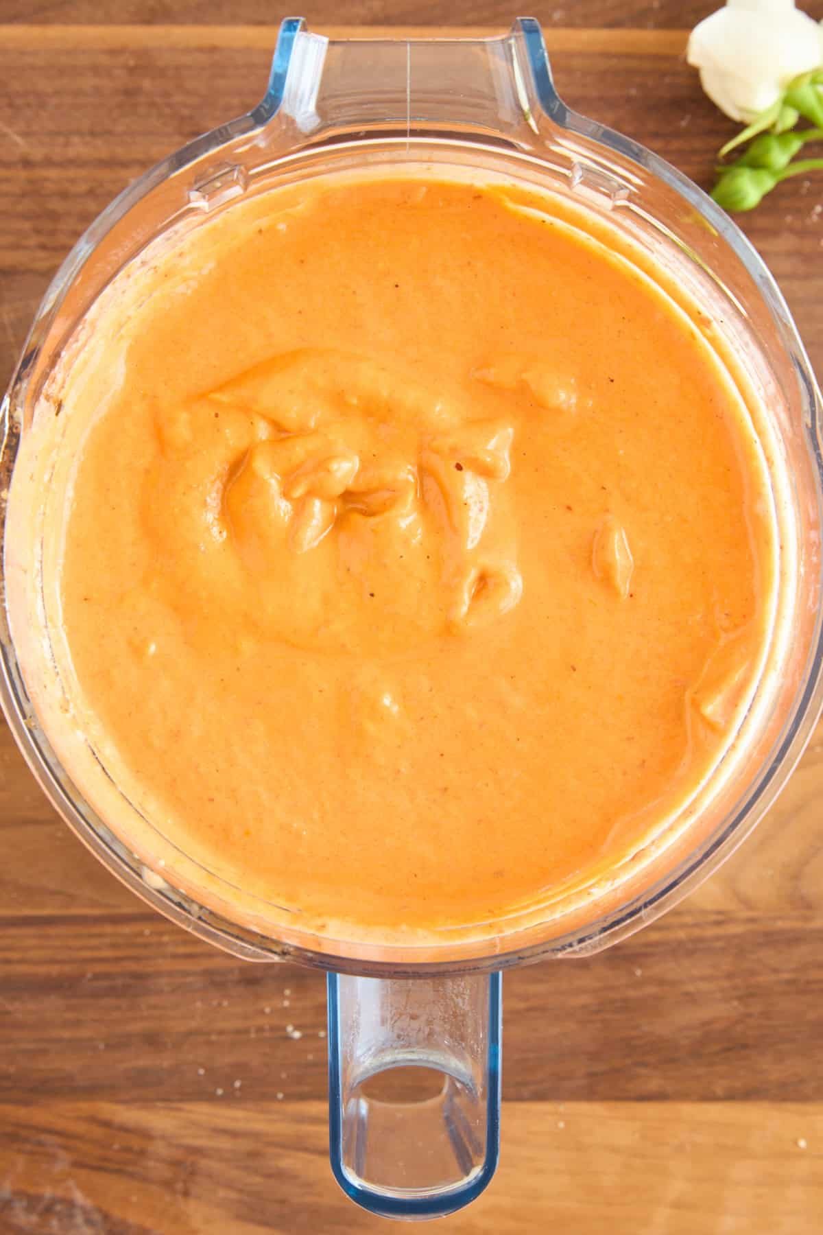 A blender filled with creamy, orange-colored sauce sits on a wooden surface. A small white flower and green leaves appear in the upper right corner.