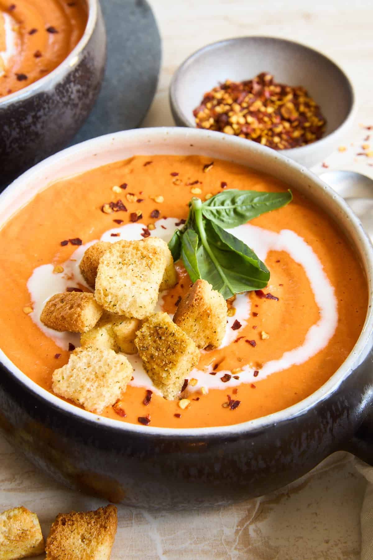 A bowl of creamy tomato soup topped with croutons, a swirl of cream, fresh basil leaves, and a sprinkle of red pepper flakes, with a small dish of red pepper flakes in the background.