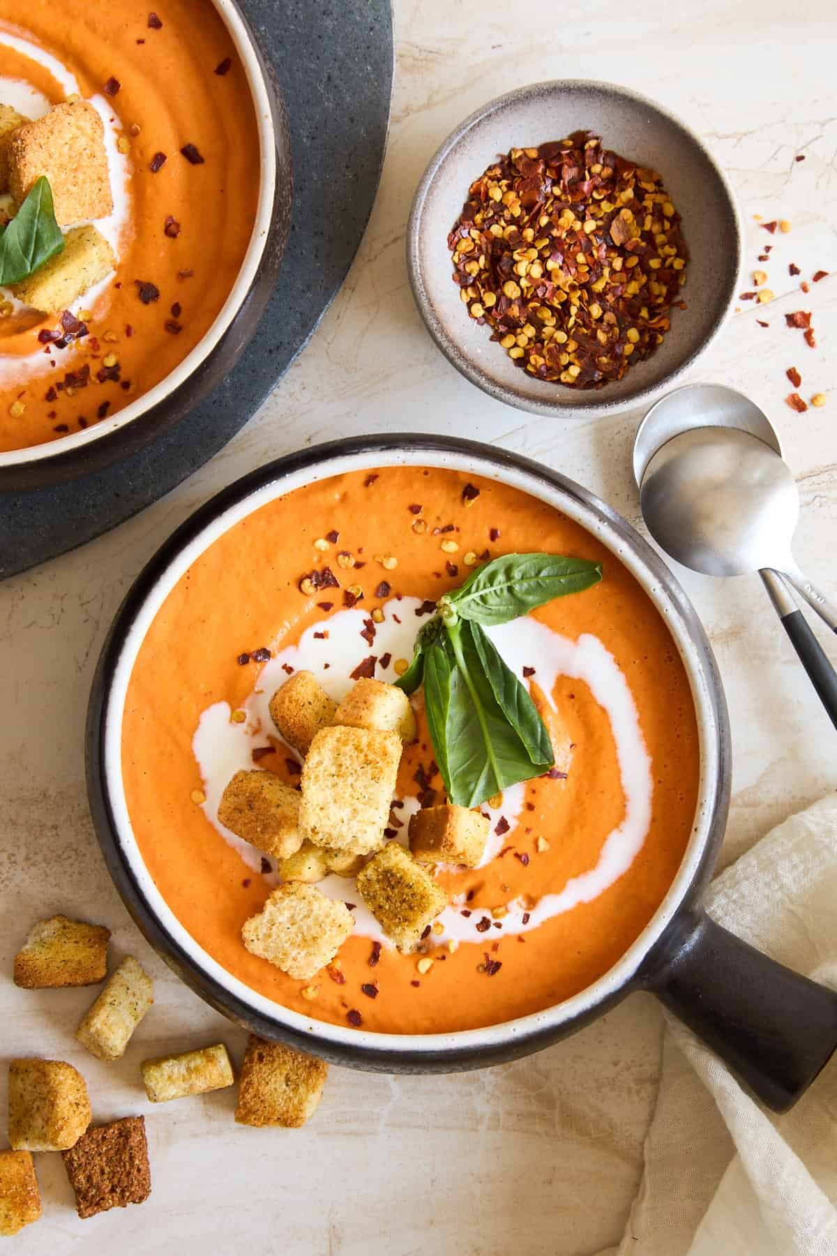 A bowl of silky tomato soup topped with croutons, fresh basil, and a swirl of cream, beside a bowl of red pepper flakes and scattered croutons, with a spoon and napkin on a light surface.