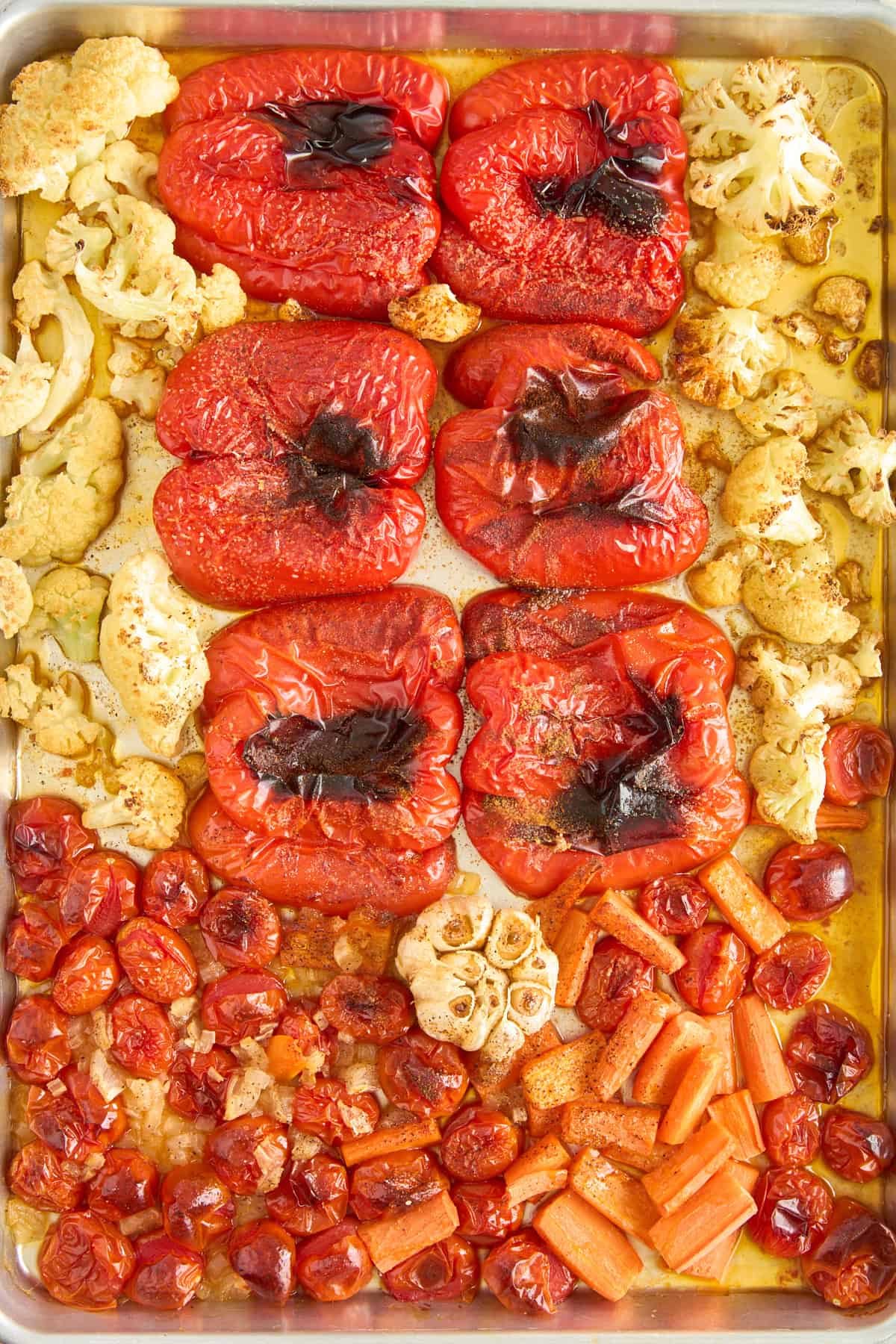 A baking sheet with roasted red bell peppers, cauliflower florets, cherry tomatoes, whole garlic bulbs, and chopped carrots, all glistening with oil and slightly charred from roasting.