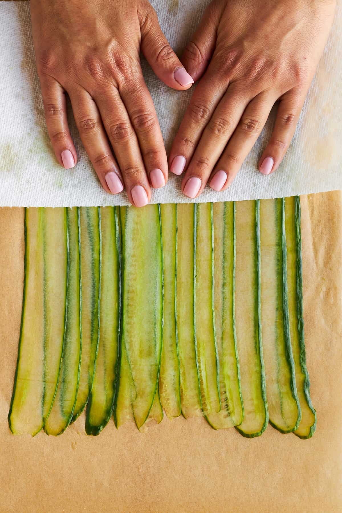 Hands patting cucumber strips dry with a paper towel.