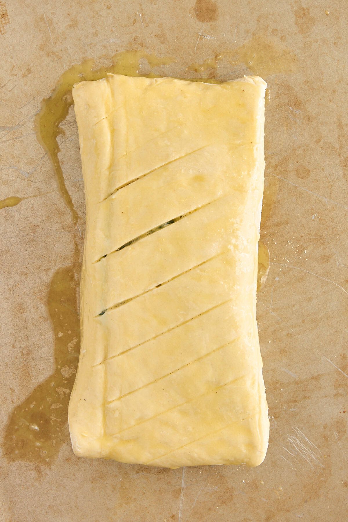 A rectangular pastry wrapped in dough with diagonal slits on top, placed on a lightly stained baking sheet. The dough appears brushed with egg wash and is ready to be baked.