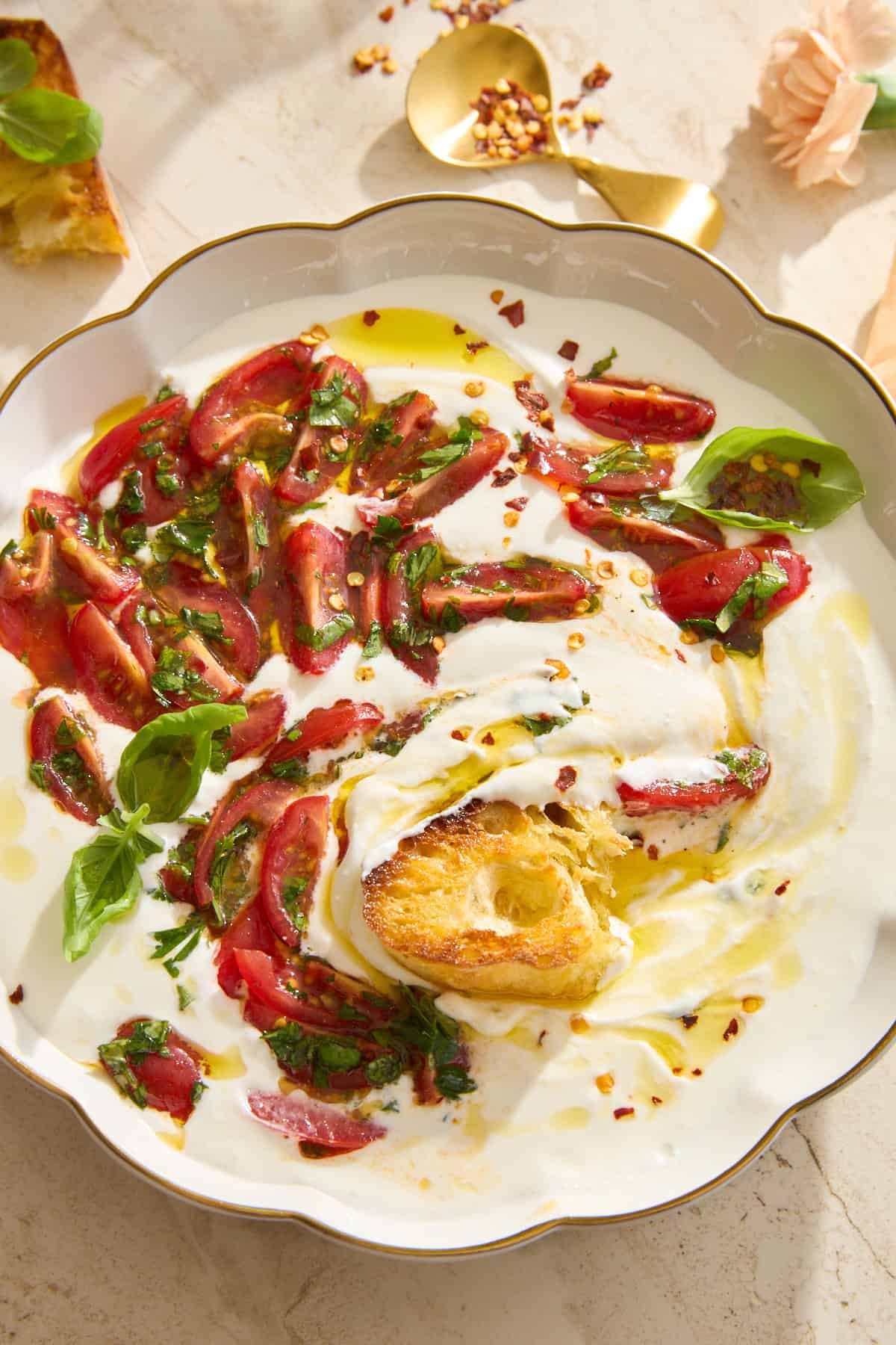 A white plate holds whipped cottage cheese topped with sliced tomatoes, fresh basil, olive oil, and red pepper flakes. A toasted bread slice is dipped into the cottage cheese, and a gold spoon rests nearby on a light surface.