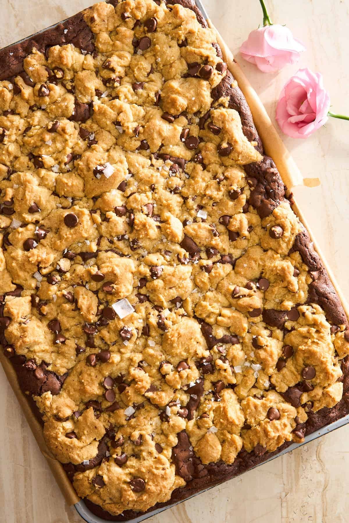 A large tray of chocolate chip cookie bars with a golden, crumbly topping and chocolate chips, set on parchment paper with two pink flowers nearby on a light surface.