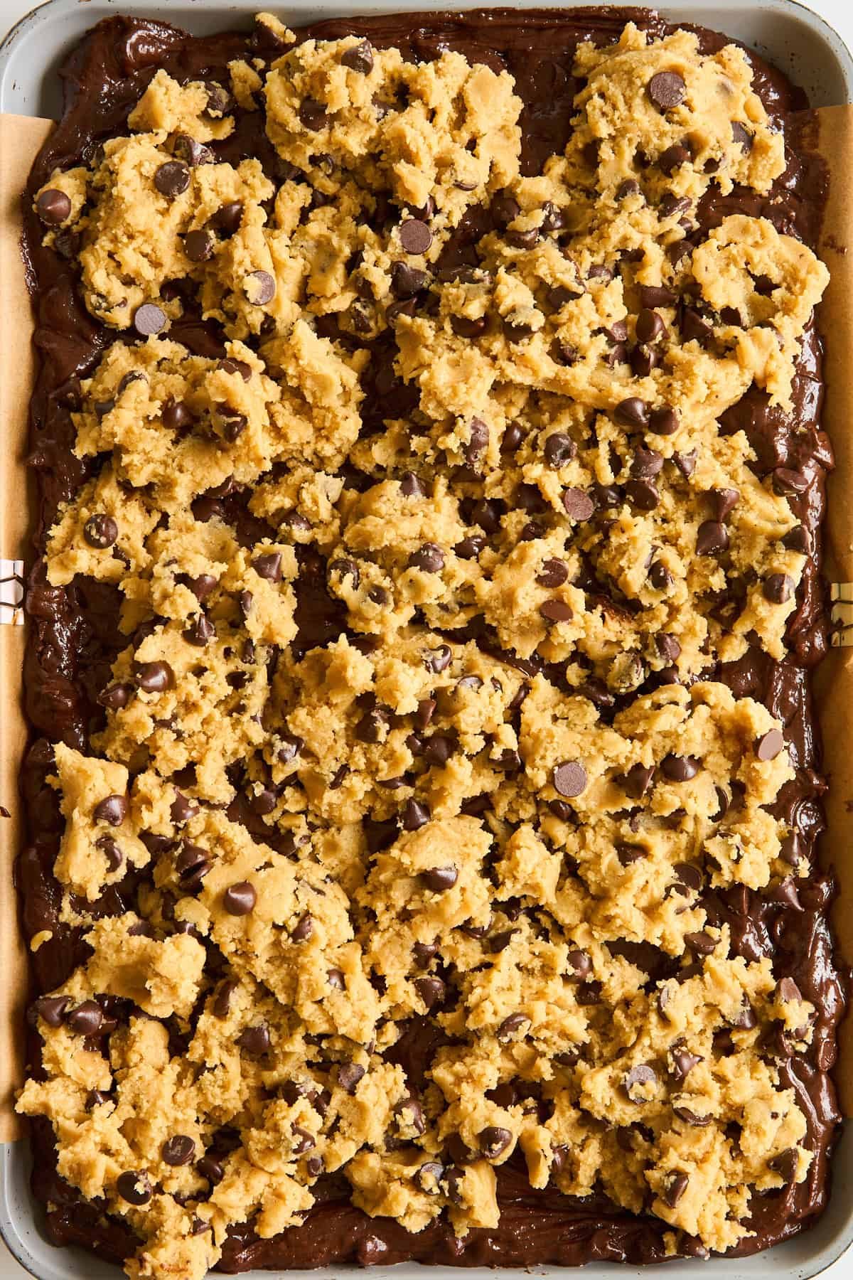 A baking tray filled with brownie batter topped with uneven scoops of chocolate chip cookie dough, ready to be baked.