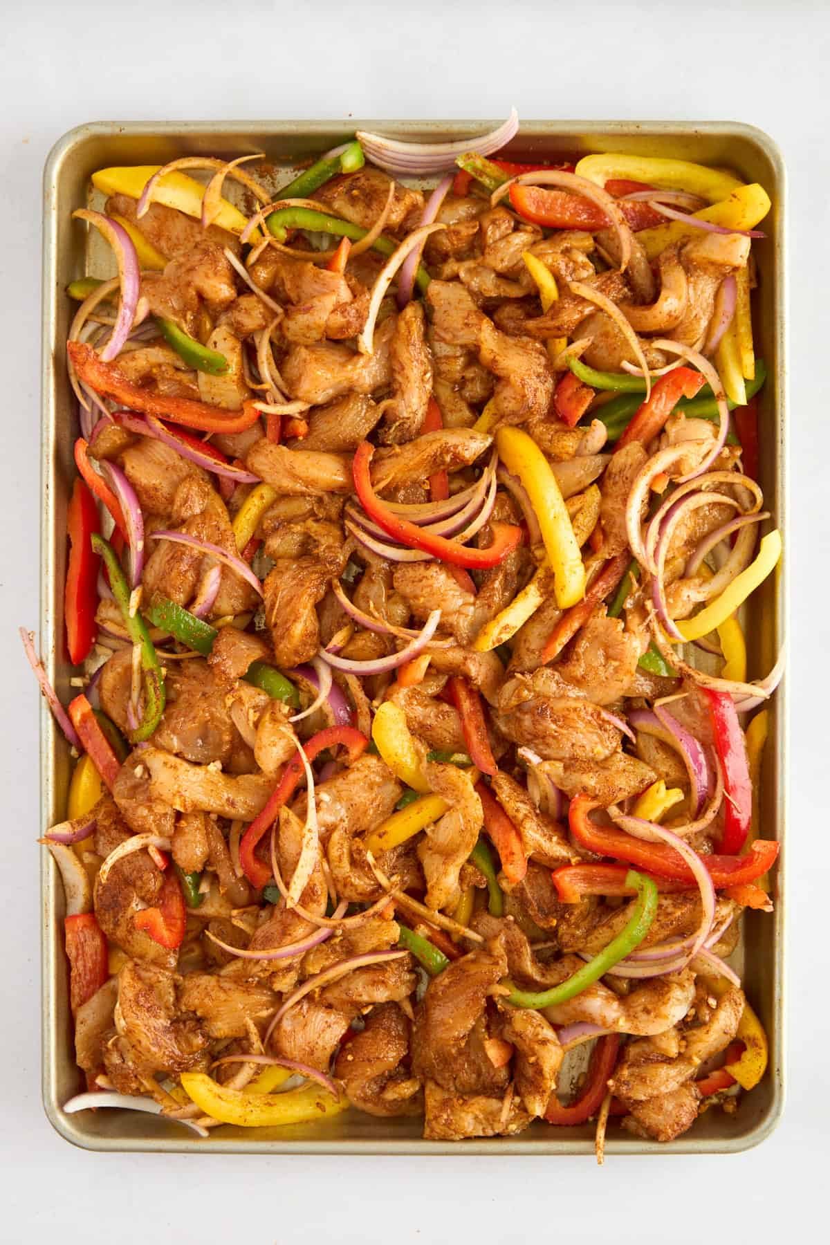 Unbaked fajita seasoned chicken slices, bell peppers, and red onions on a baking sheet.