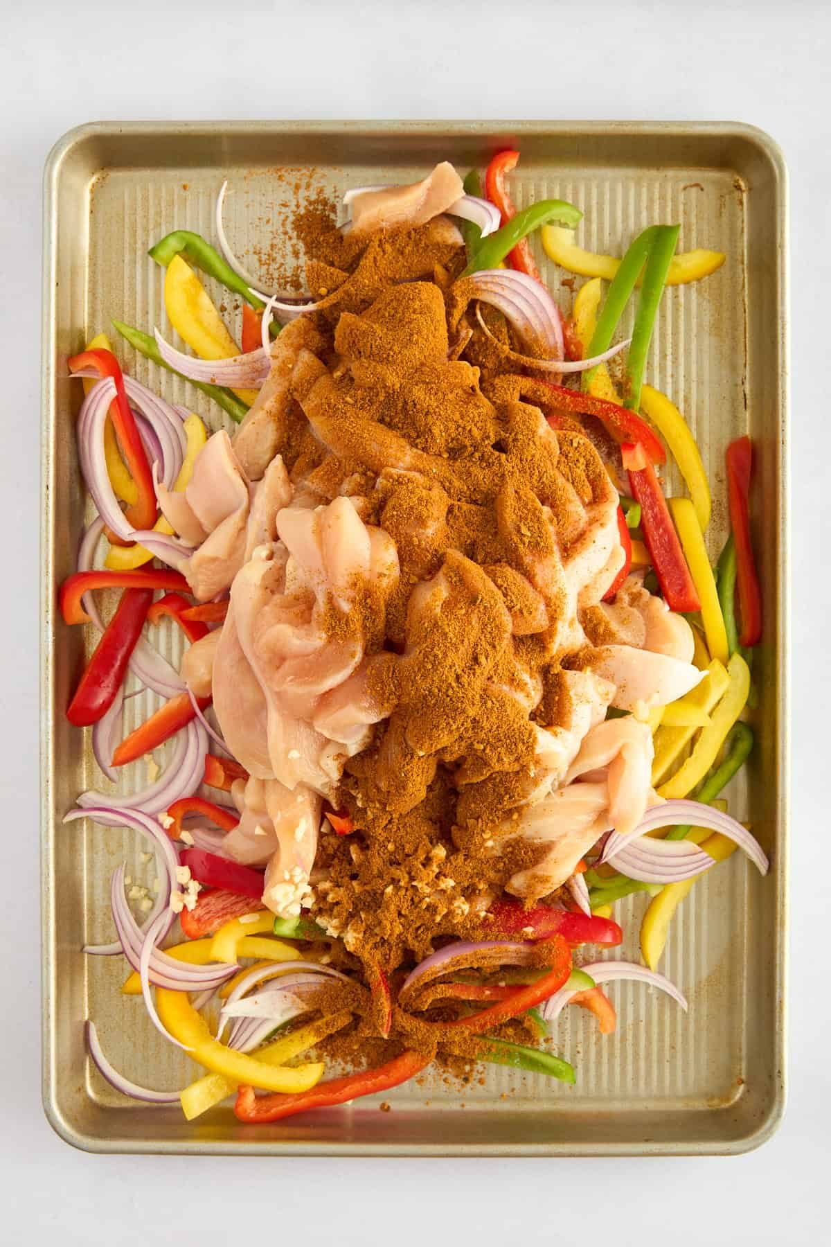 Raw chicken breasts, sliced red onions, and sliced peppers on a baking sheet topped with fajita seasoning.