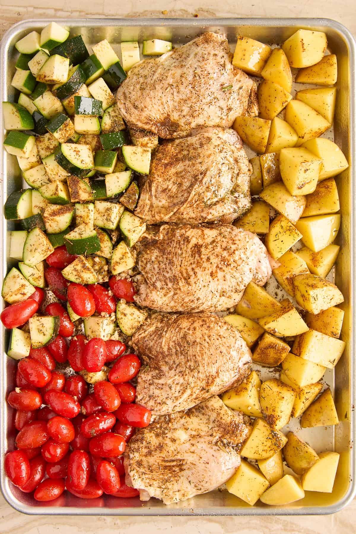 A baking sheet with seasoned chicken thighs in the center, surrounded by chopped zucchini, halved cherry tomatoes, and diced potatoes, all sprinkled with spices, ready to be cooked.