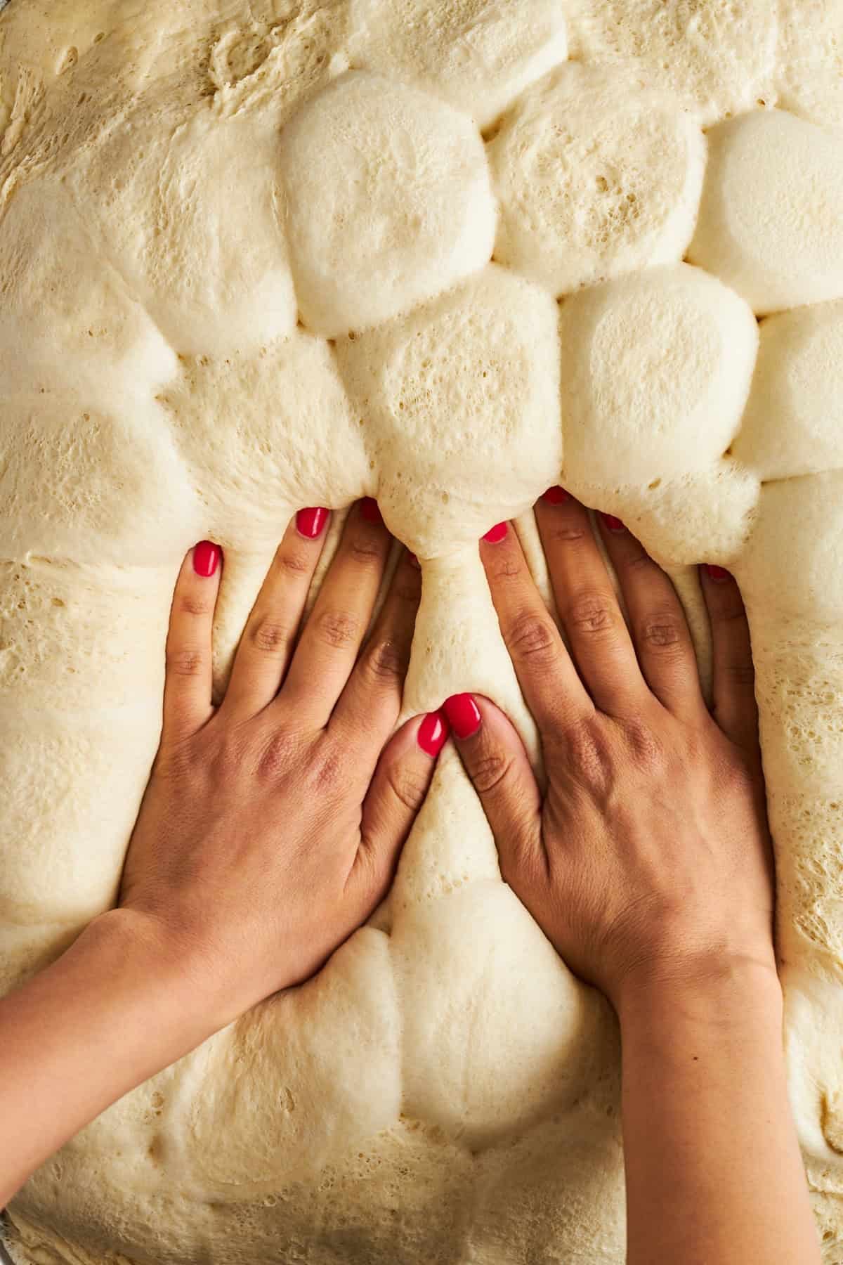 Two hands pressing down raw dinner rolls to form a flat crust.