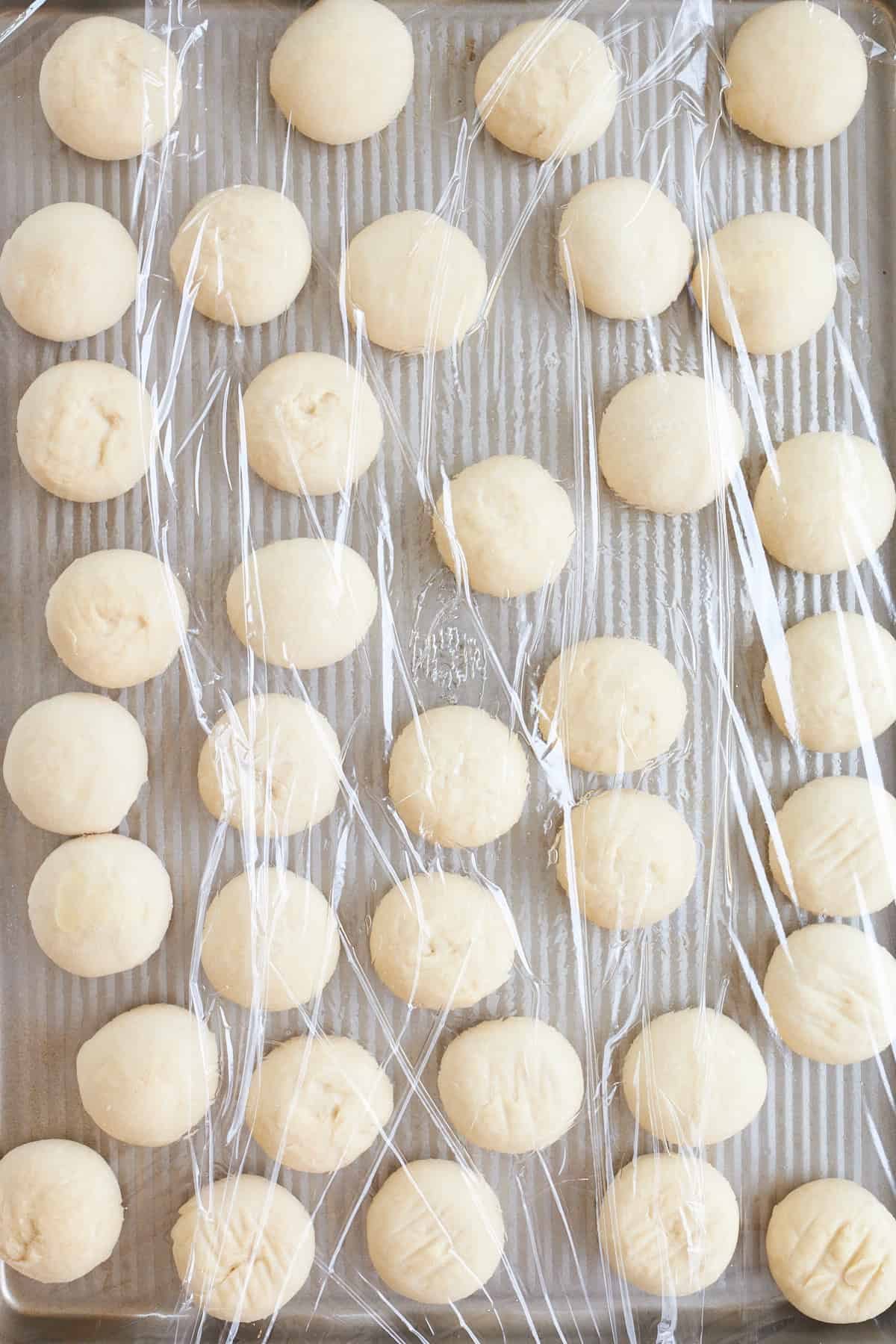 Raw dinner rolls on a baking sheet covered with plastic wrap.