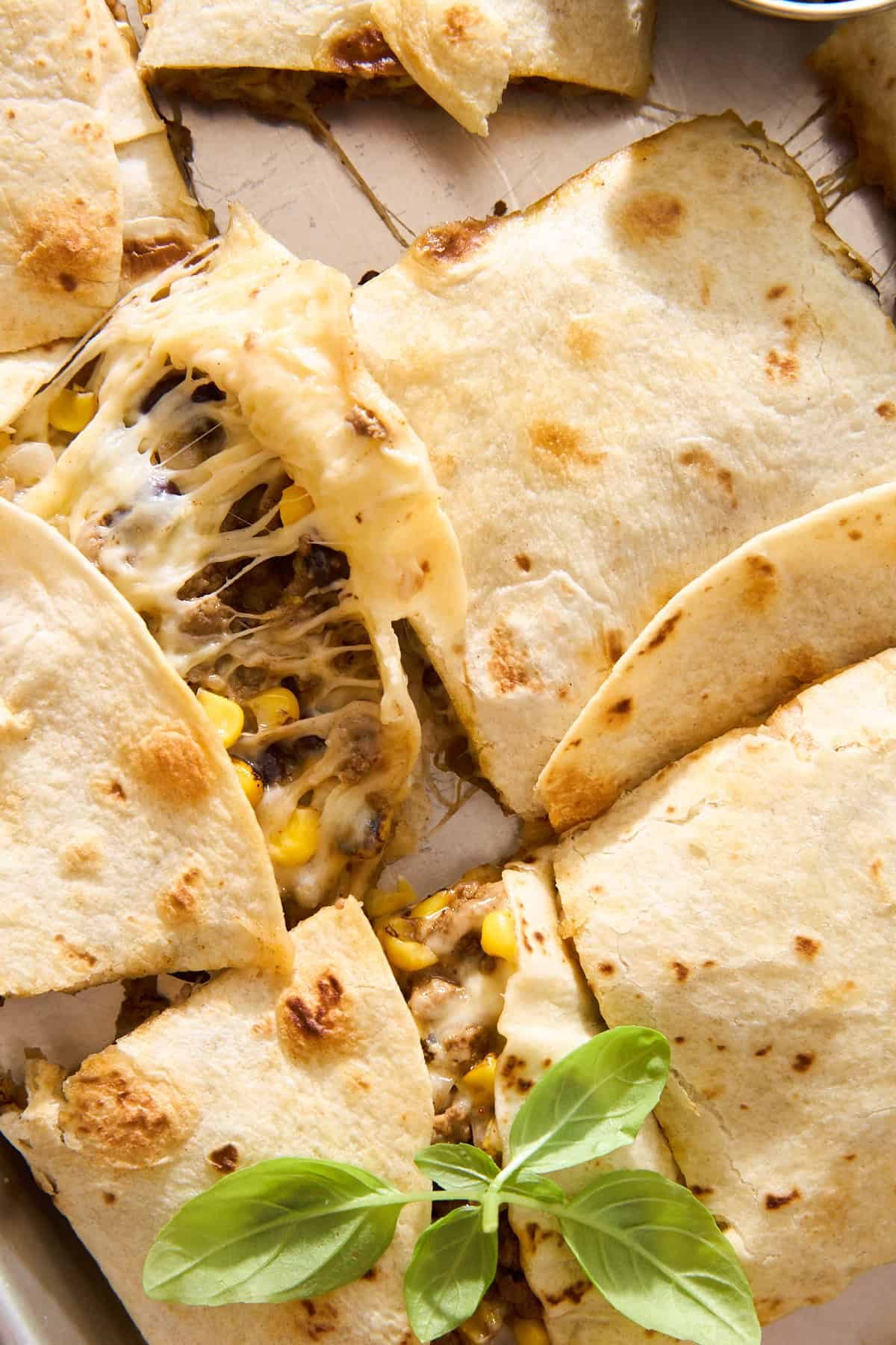 A close-up of sliced quesadillas with melted cheese, corn, and beef filling on a tray, garnished with fresh basil leaves. One slice is being pulled, showing the gooey cheese stretch.