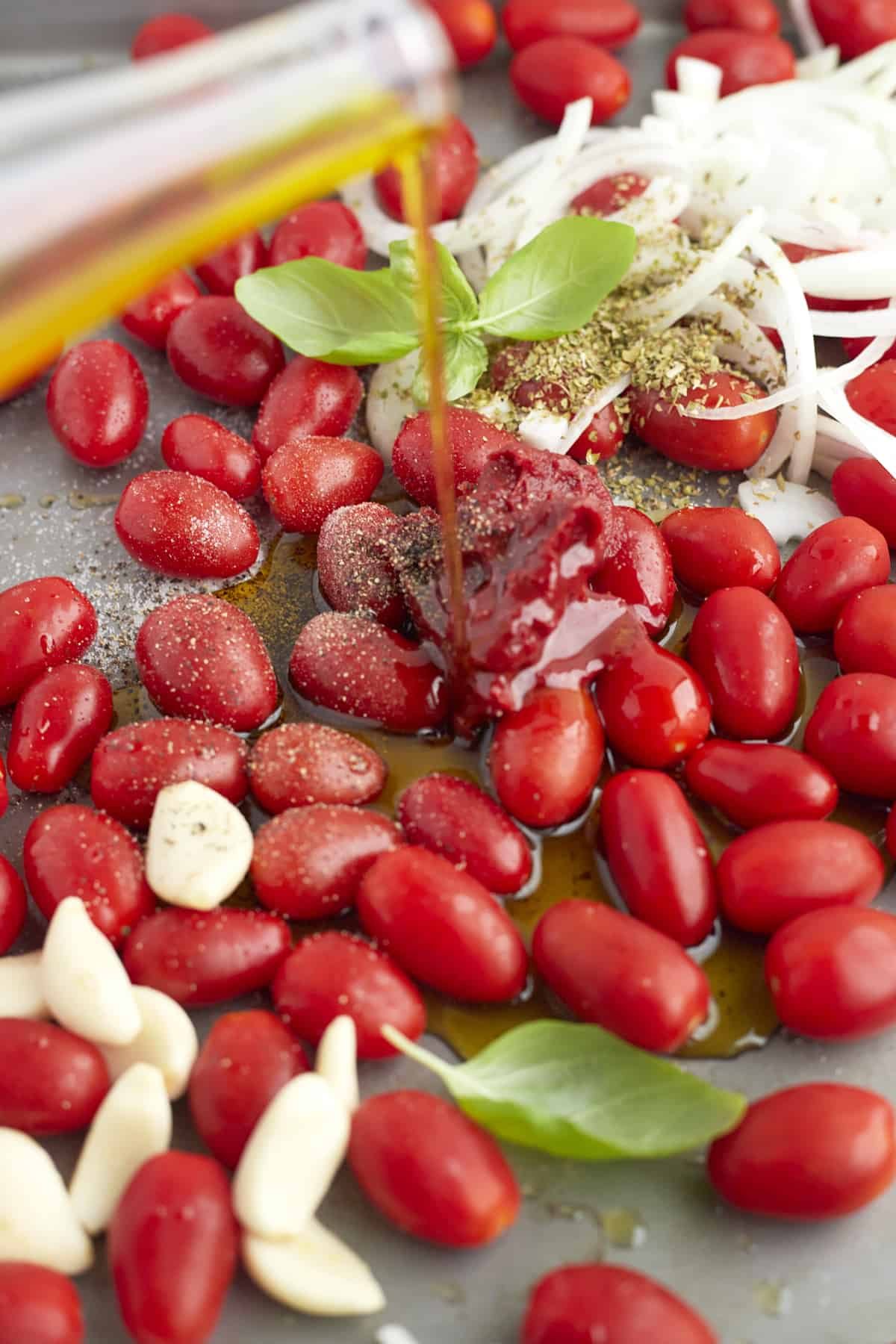 garlic cloves, tomatoes, and onions on a baking sheet topped with seasonings with olive oil being drizzled on top