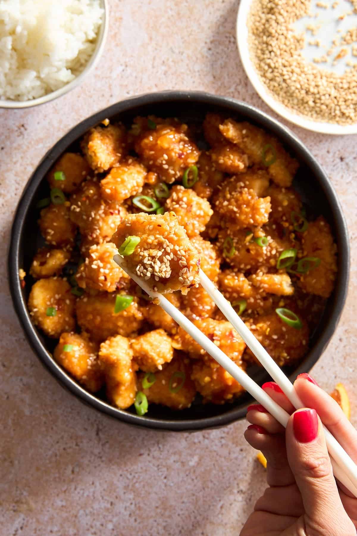 Two chopsticks holding a piece of orange chicken over a bowl.