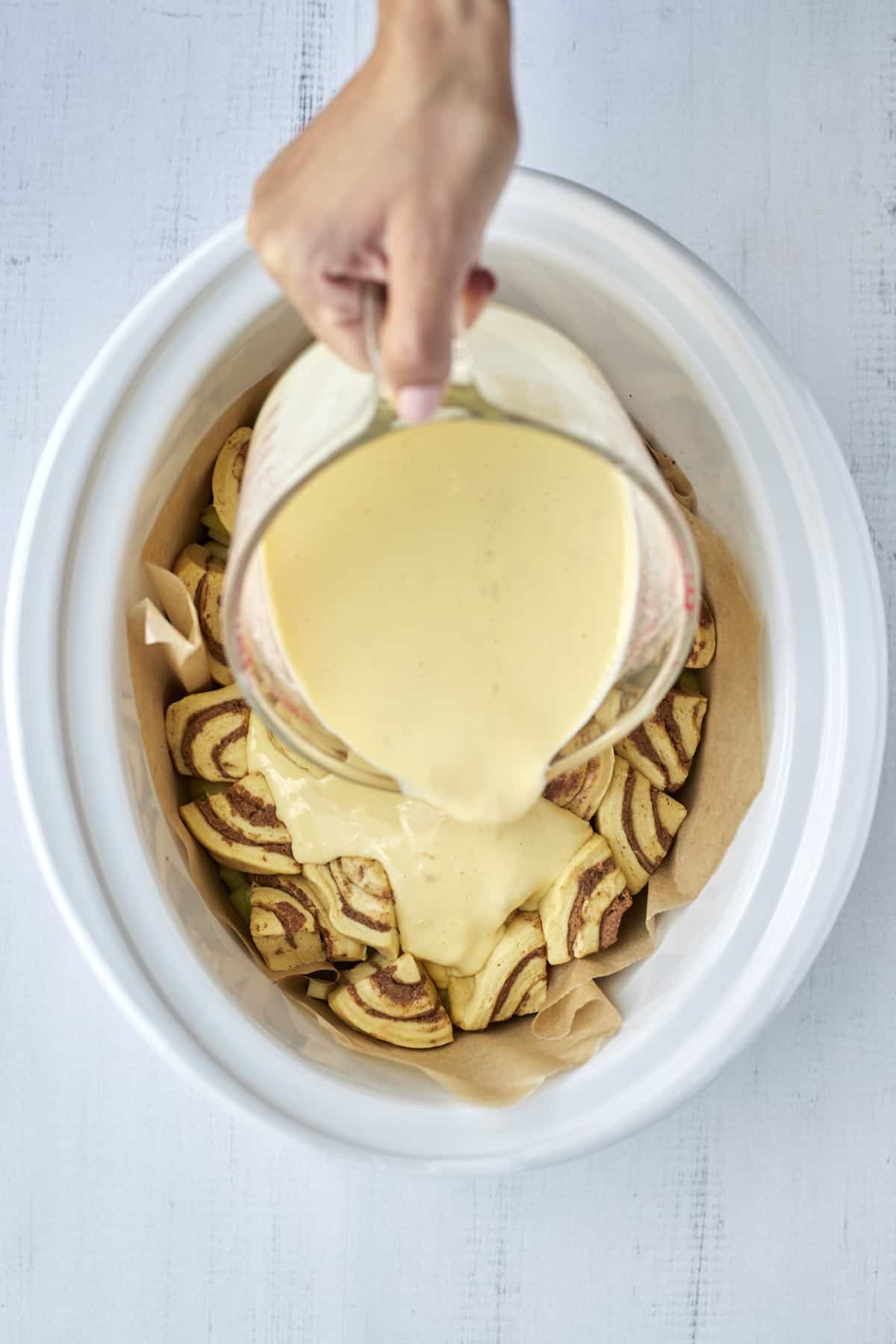 a hand pouring a custard mixture over layers of cinnamon rolls pieces and apples in a parchment lined slow cooker.