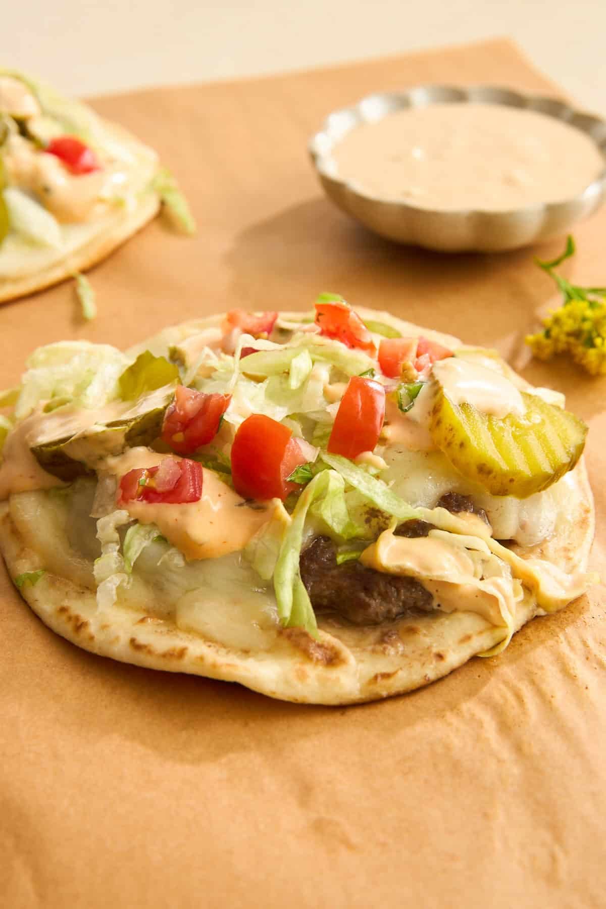 A flatbread topped with ground beef, melted cheese, shredded lettuce, diced tomatoes, pickles, and a silky sauce sits on brown parchment paper, with a small dish of extra sauce in the background.