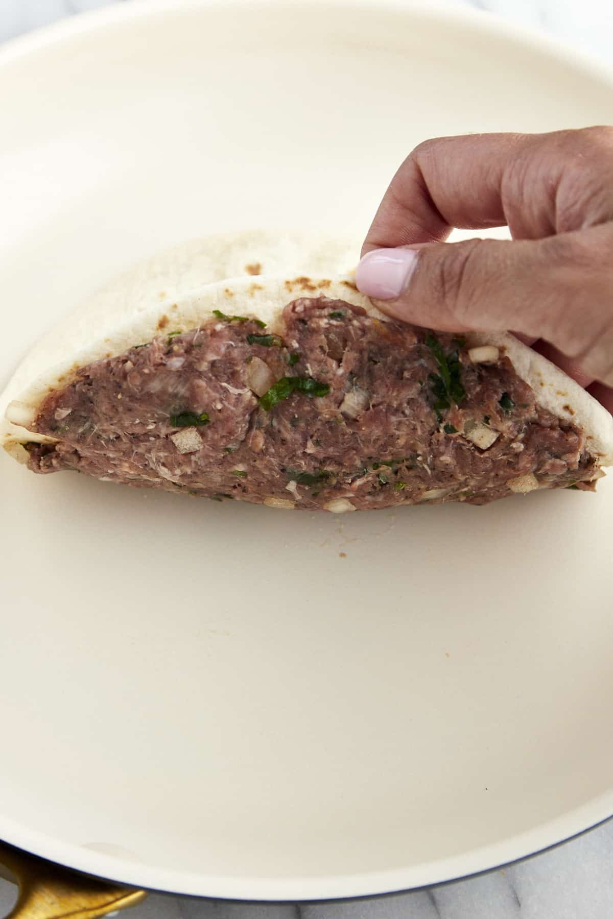 A hand placing a tortilla with seasoned ground beef on it into a pan.