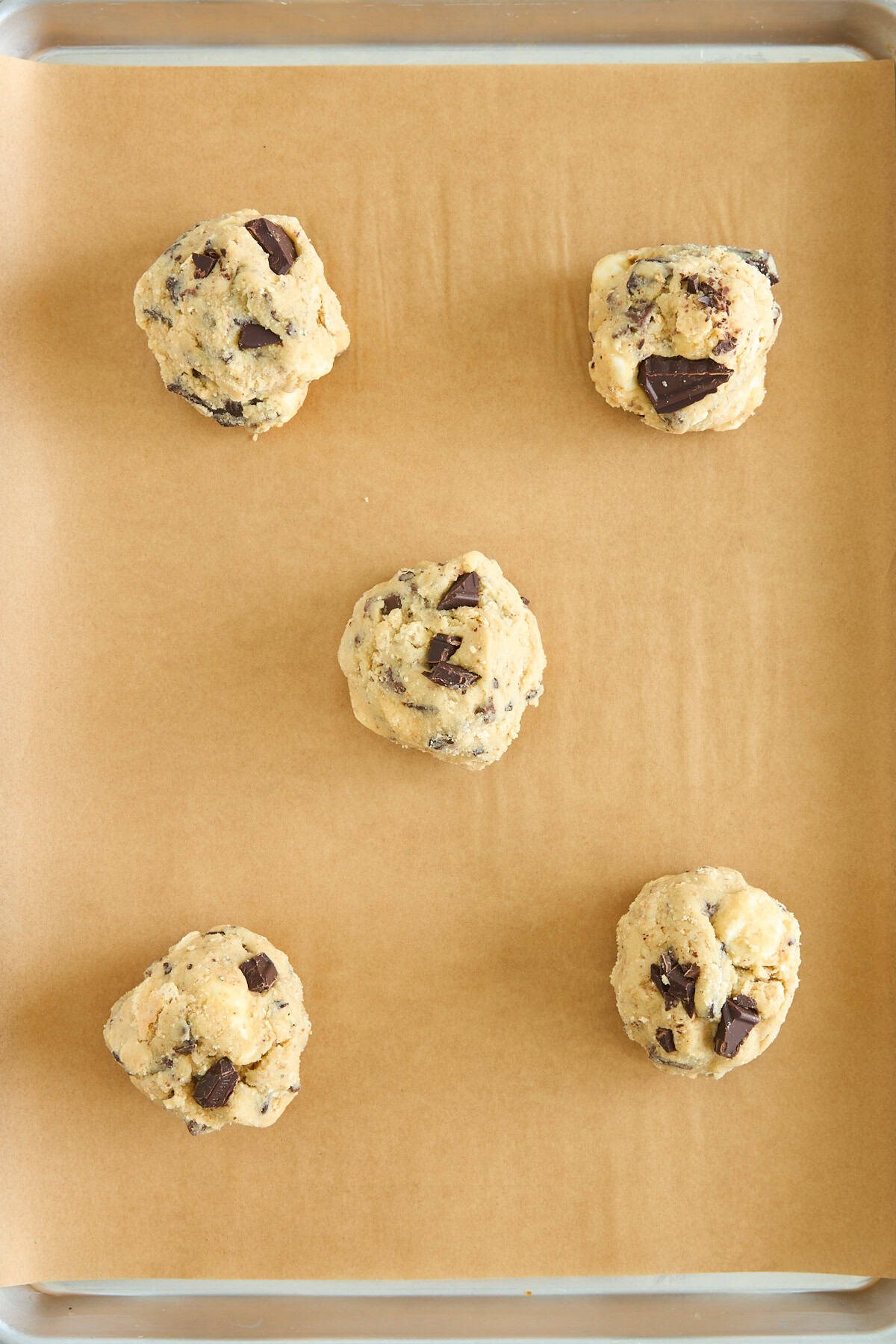 Five balls of chocolate chip cookie dough are evenly spaced on a parchment-lined baking sheet, ready to be baked.