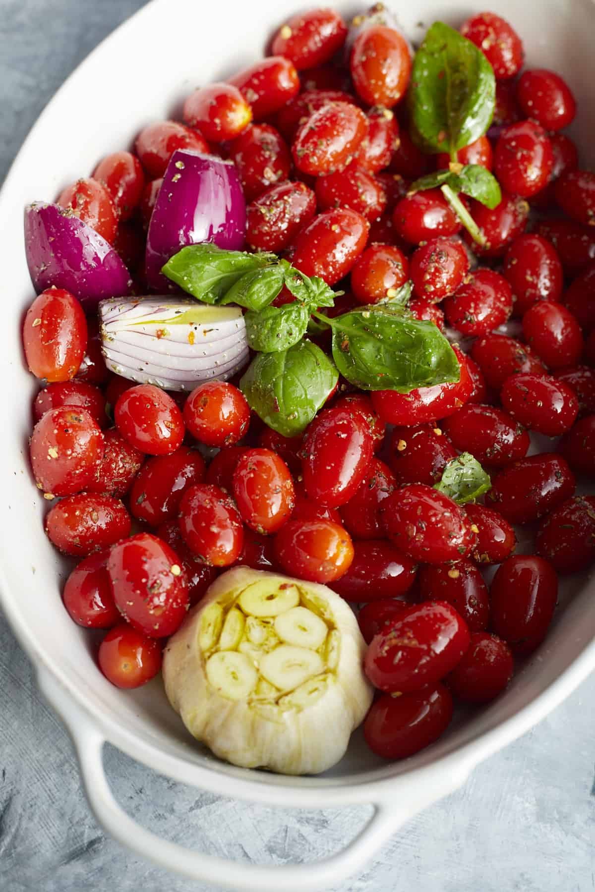 overhead image of tomatoes, garlics, shallot, onions, basil, and seasoning drizzled with olive oil in a white baking dish