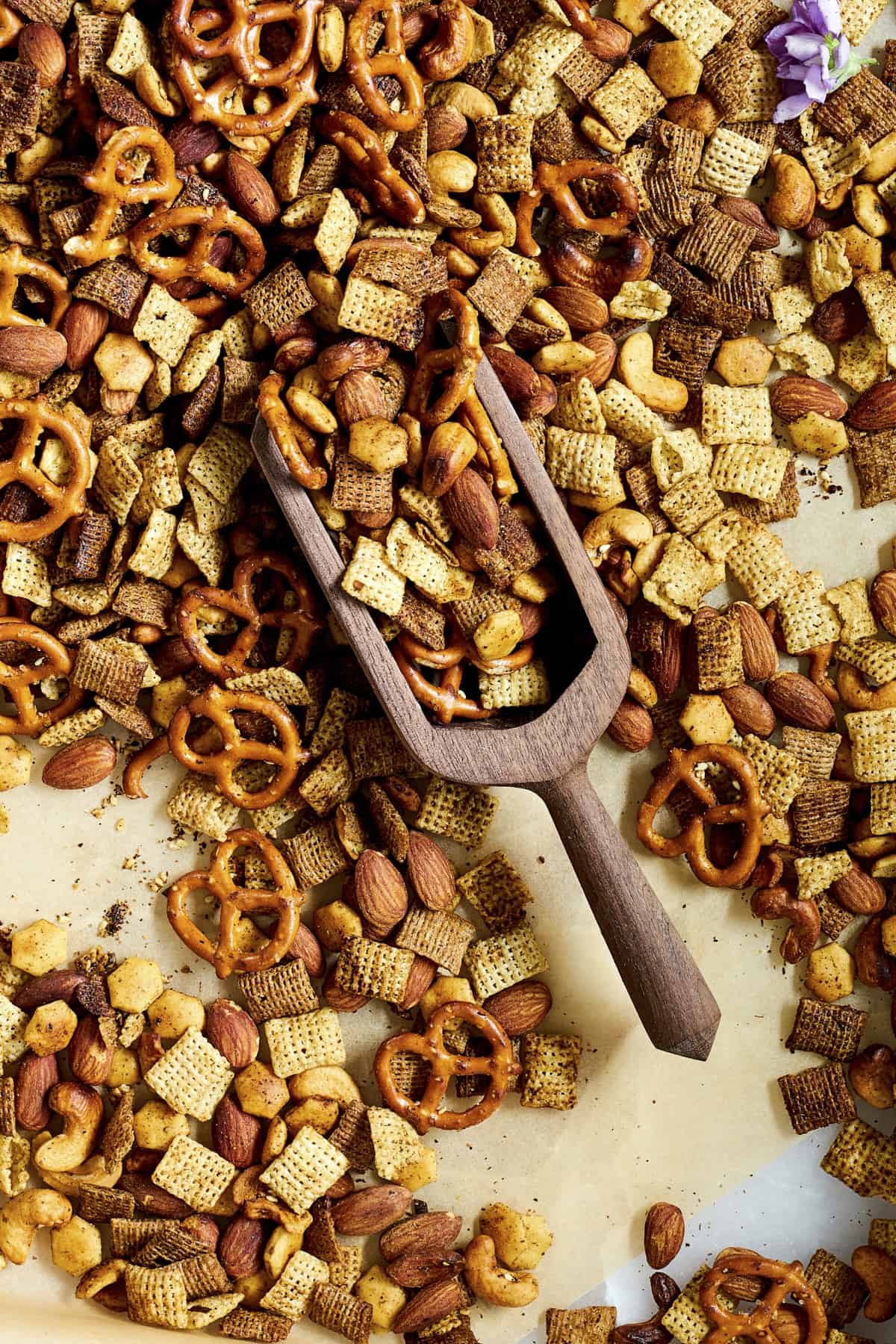A wooden scoop lifting spicy Chex mix from a baking sheet.