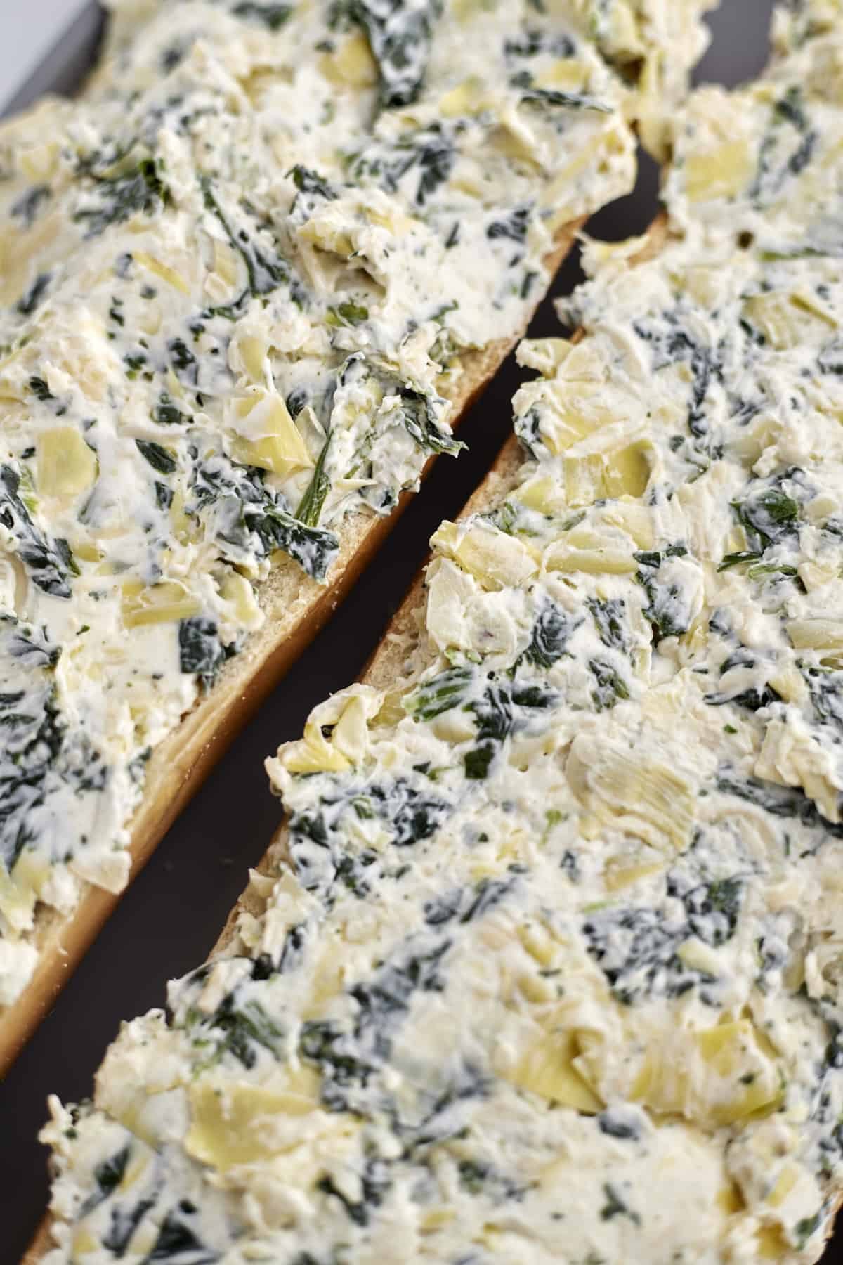 two halves of french bread topped with a spinach and artichoke mixture ready to be baked for homemade garlic bread