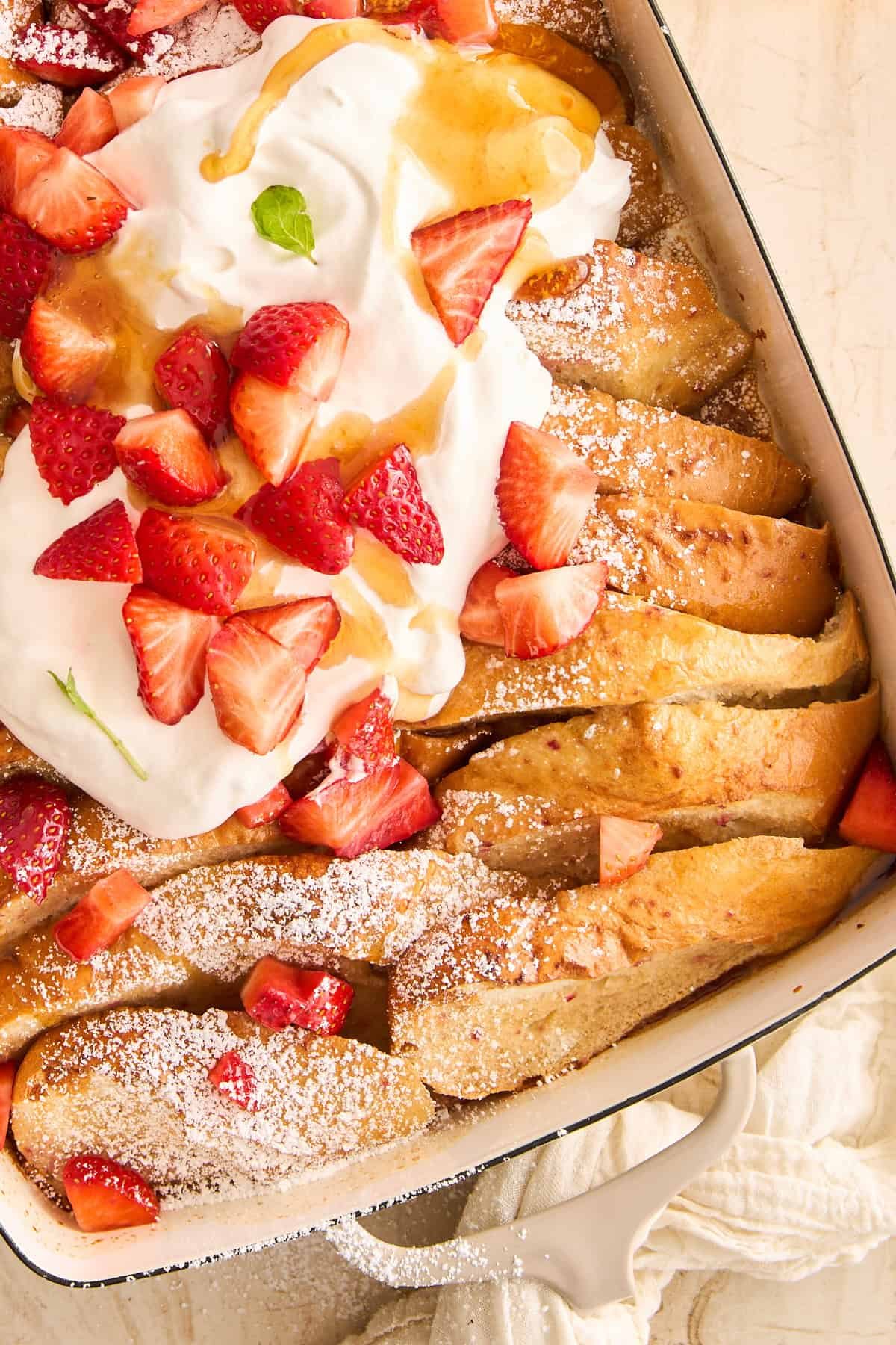 A baking dish of strawberry french toast topped with whipped cream, berries, and syrup.