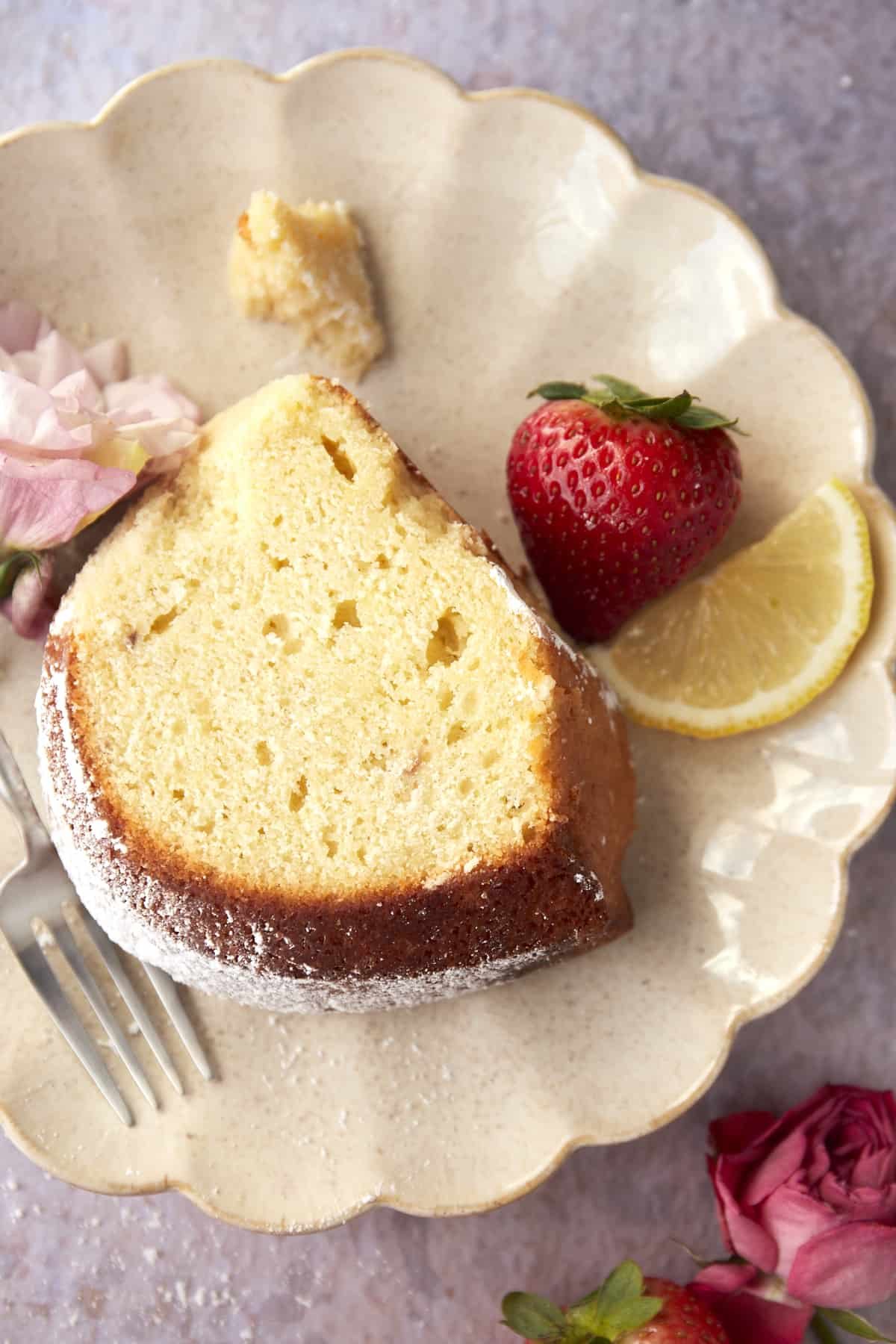 overhead image of a piece of bundt cake topped with powdered sugar on a plate with a fork, a strawberry, and a lemon slice