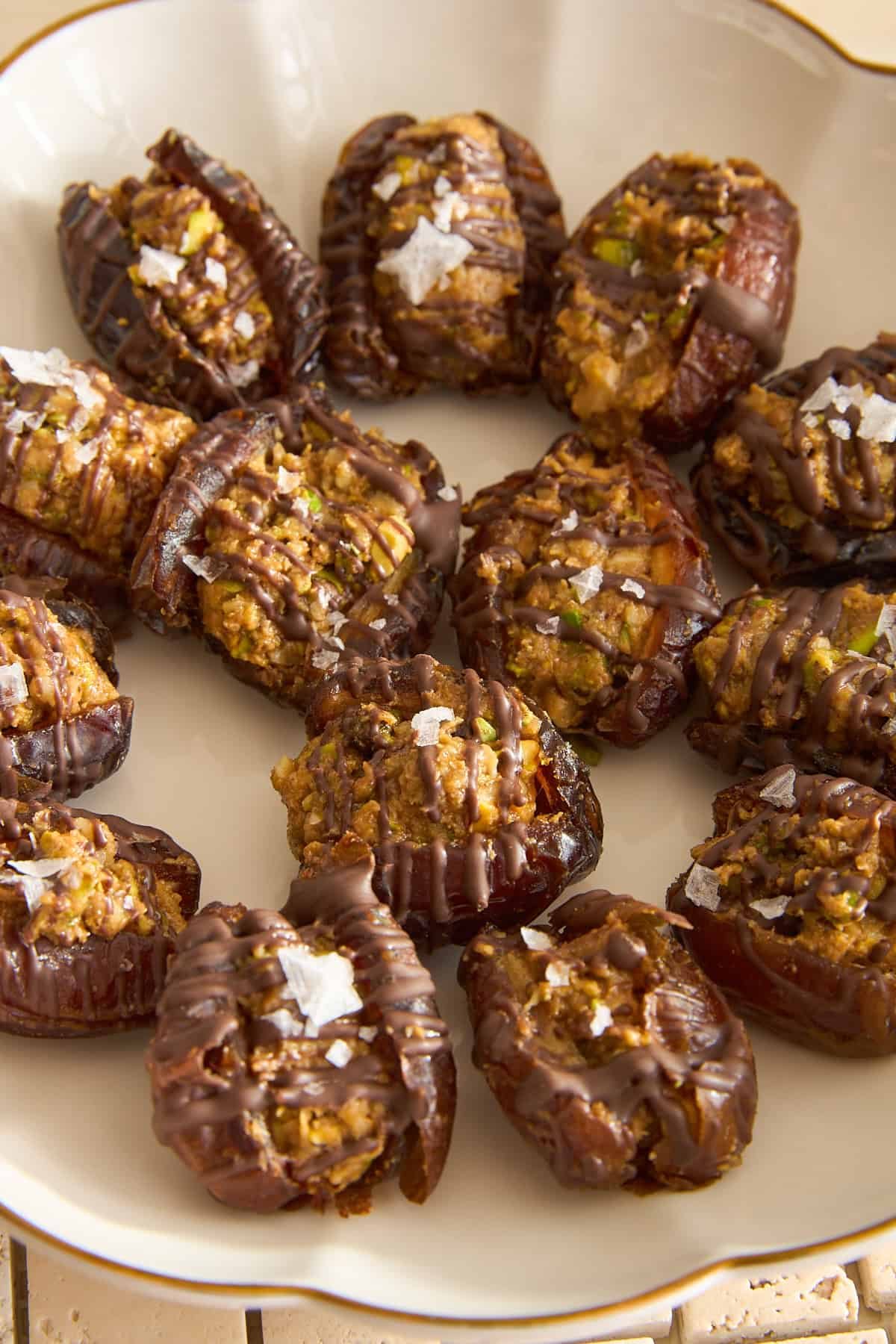 A plate of stuffed dates topped with drizzled chocolate and flakes of sea salt, arranged in a circular pattern on a cream-colored dish.