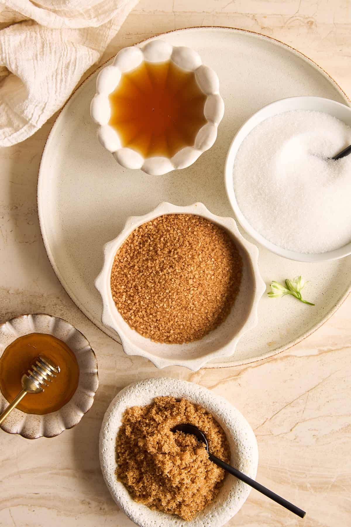 A top-down view of various sweeteners in small bowls on a beige surface, including honey with a dipper, brown sugar, white sugar, and a golden liquid, each with a spoon.