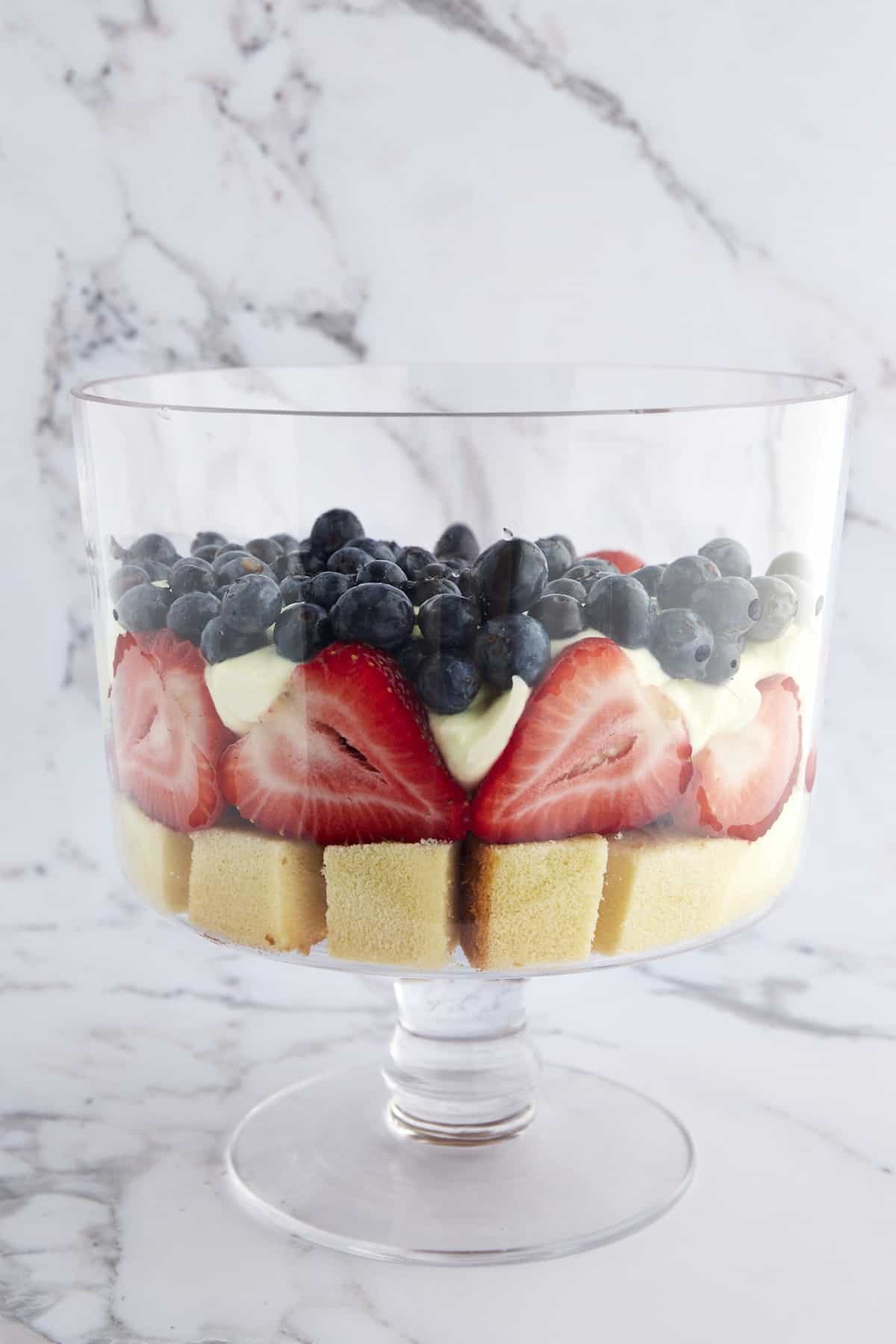 A trifle bowl with a layer of pound cake, strawberries, whipped topping, and blueberries.