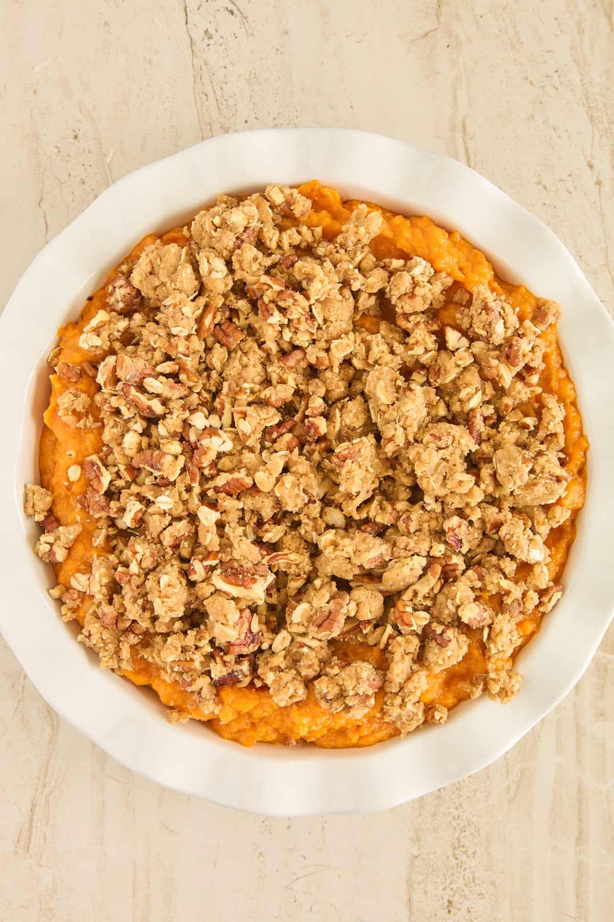 A white pie dish filled with sweet potato casserole topped with a crumbly pecan and oat streusel, on a light-colored surface.