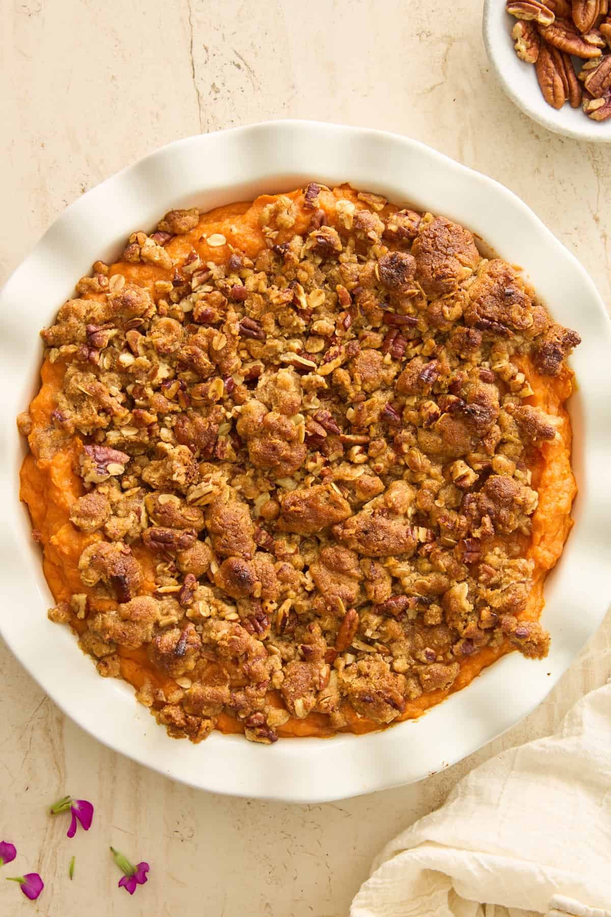 A round white dish filled with sweet potato casserole, topped with a golden-brown pecan and oat crumble. The dish sits on a light-colored surface, with a bowl of pecans and flower petals nearby.
