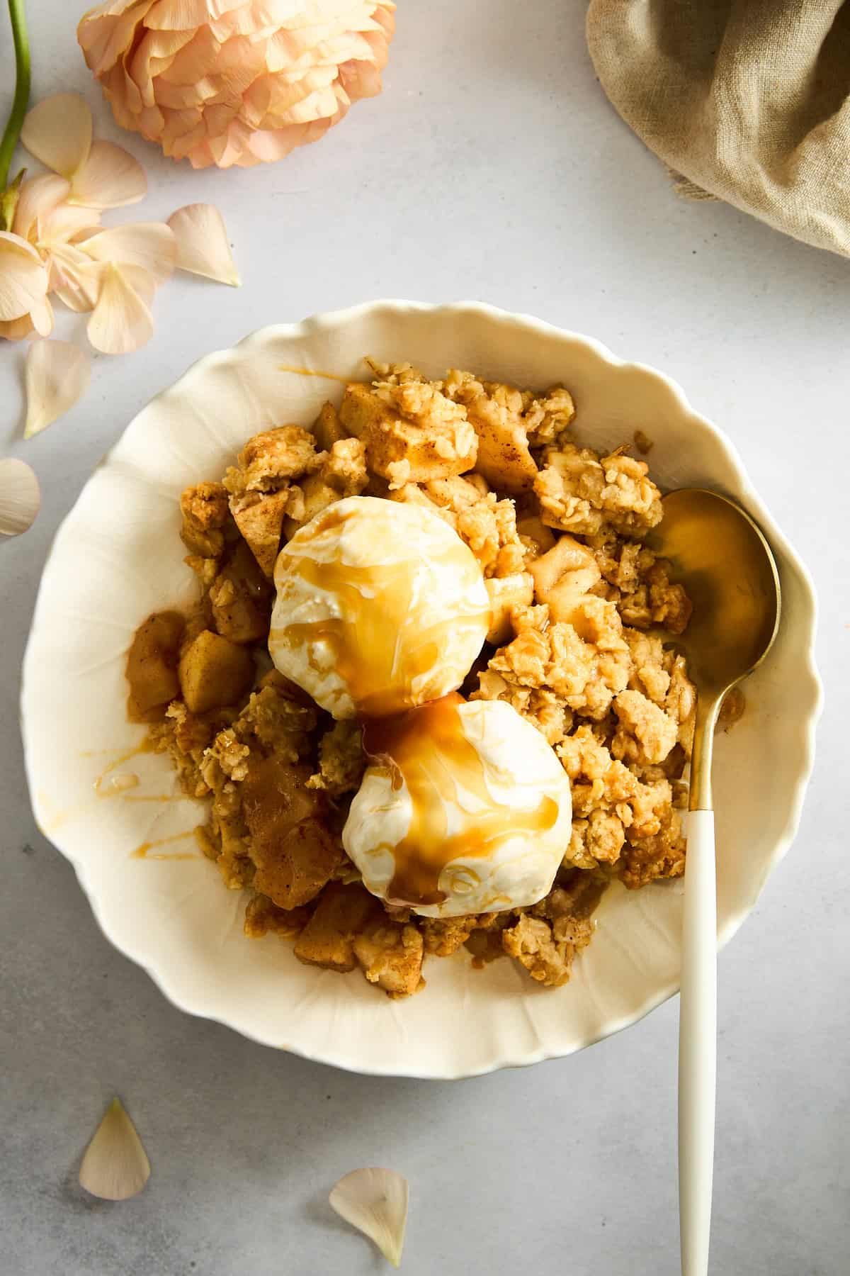 A bowl of apple crisp topped with ice cream and caramel sauce with a spoon on the side.