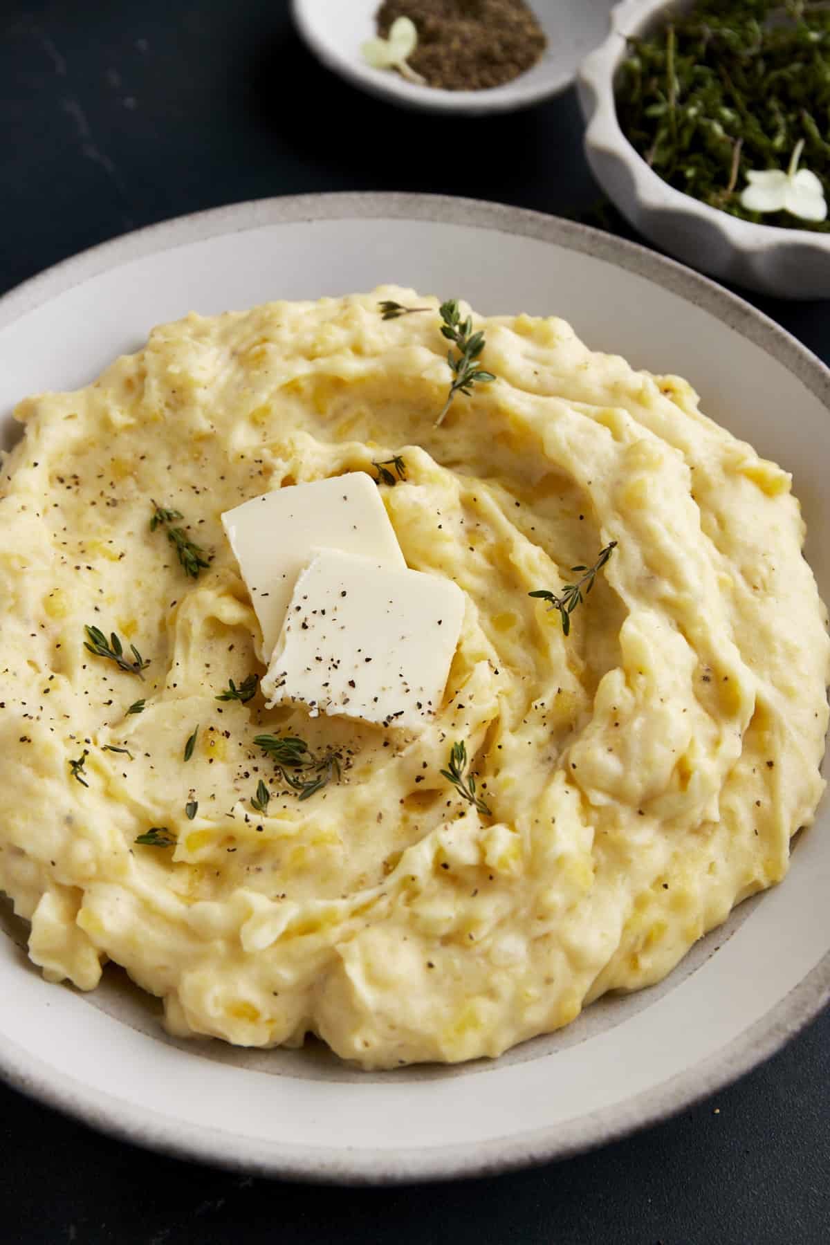 A bowl of the best mashed potatoes topped with butter, pepper, and herbs.