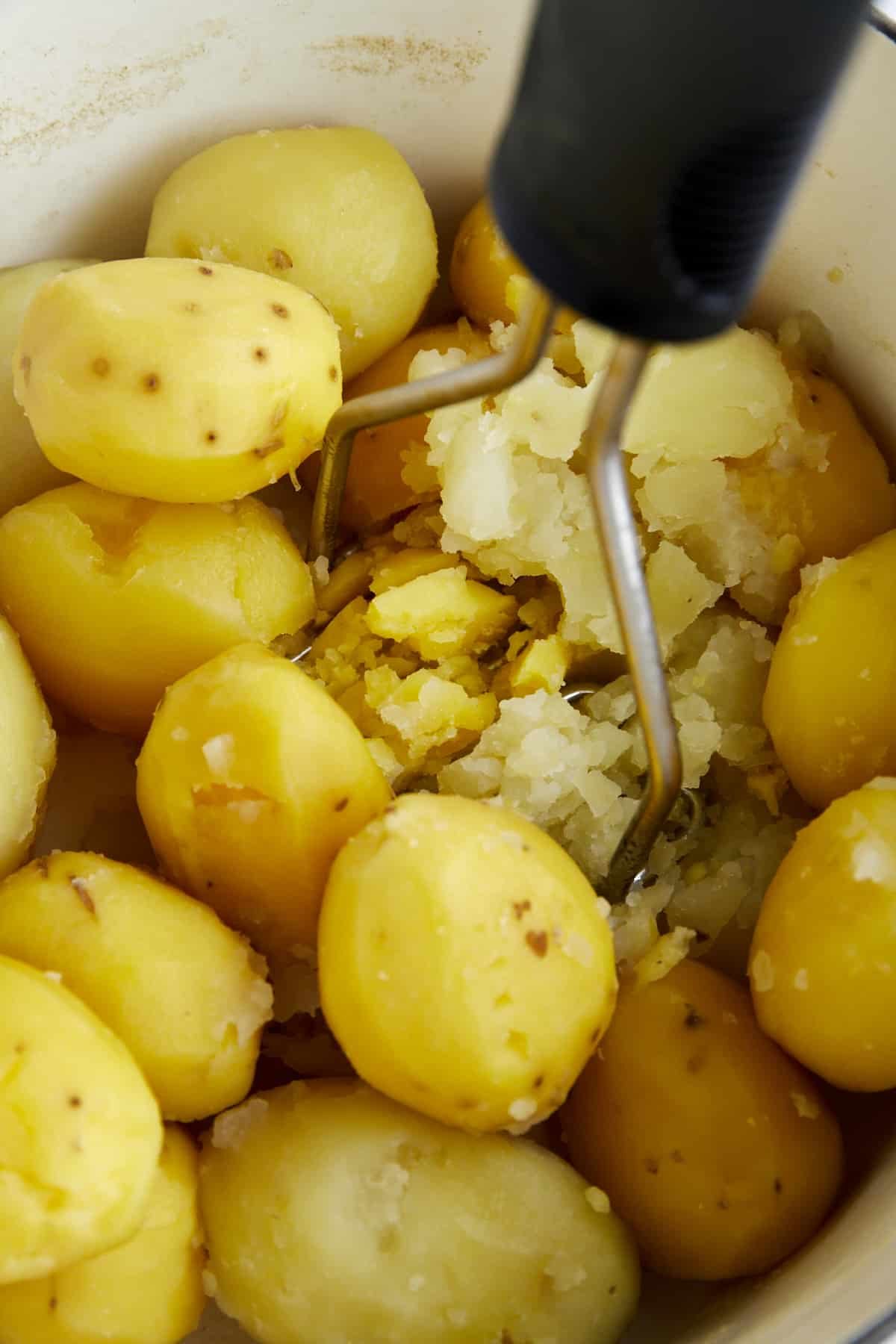 Boiled potatoes being mashed with a potato masher in a pot.