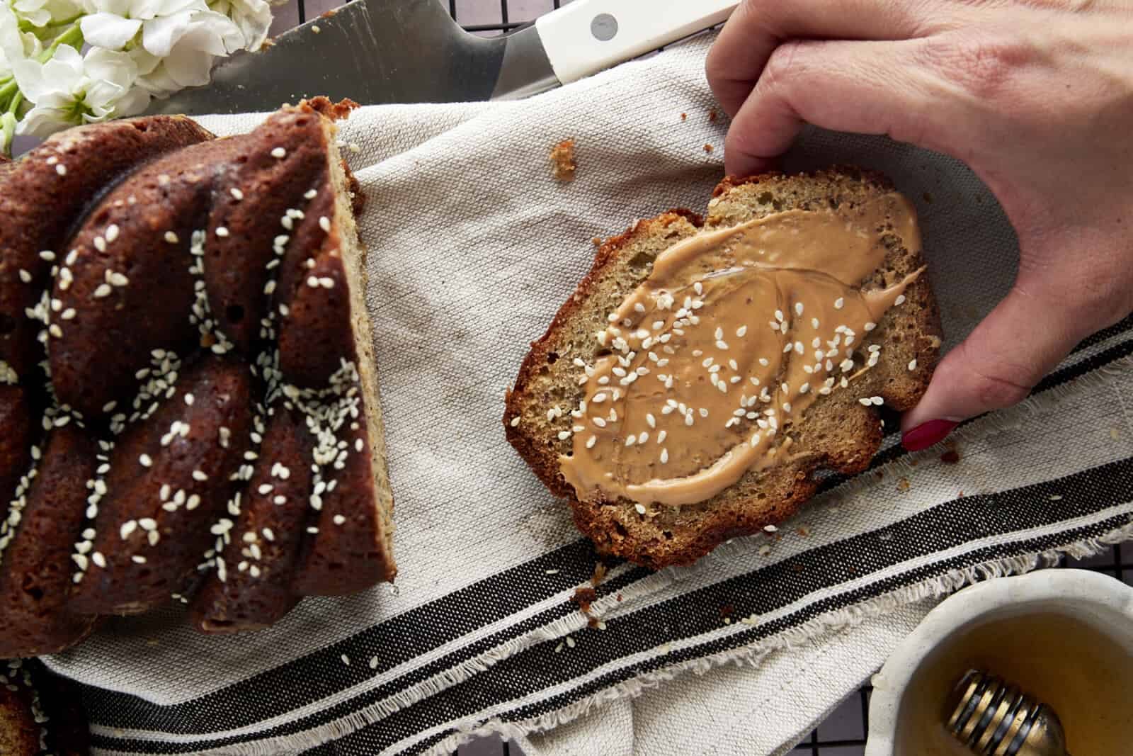 A hand grabbing a slice of tahini banana bread topped with nut butter, honey, and white sesame seeds.