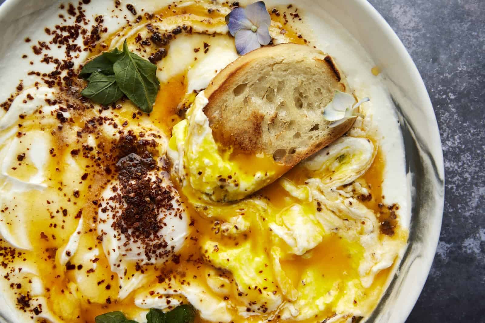 A piece of toasted bread dipping into a bowl of Turkish poached eggs with cottage cheese.