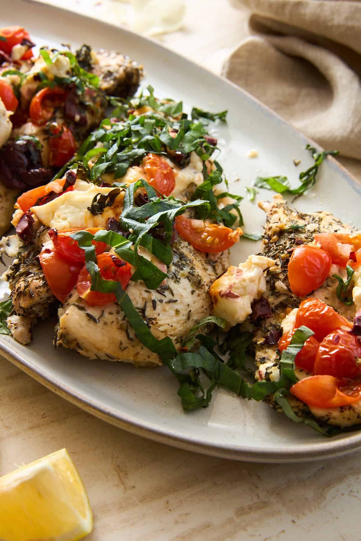 Herb-seasoned baked chicken breasts topped with chopped tomatoes, olives, feta cheese, and fresh basil, served on a white oval plate, with a lemon wedge visible in the corner.