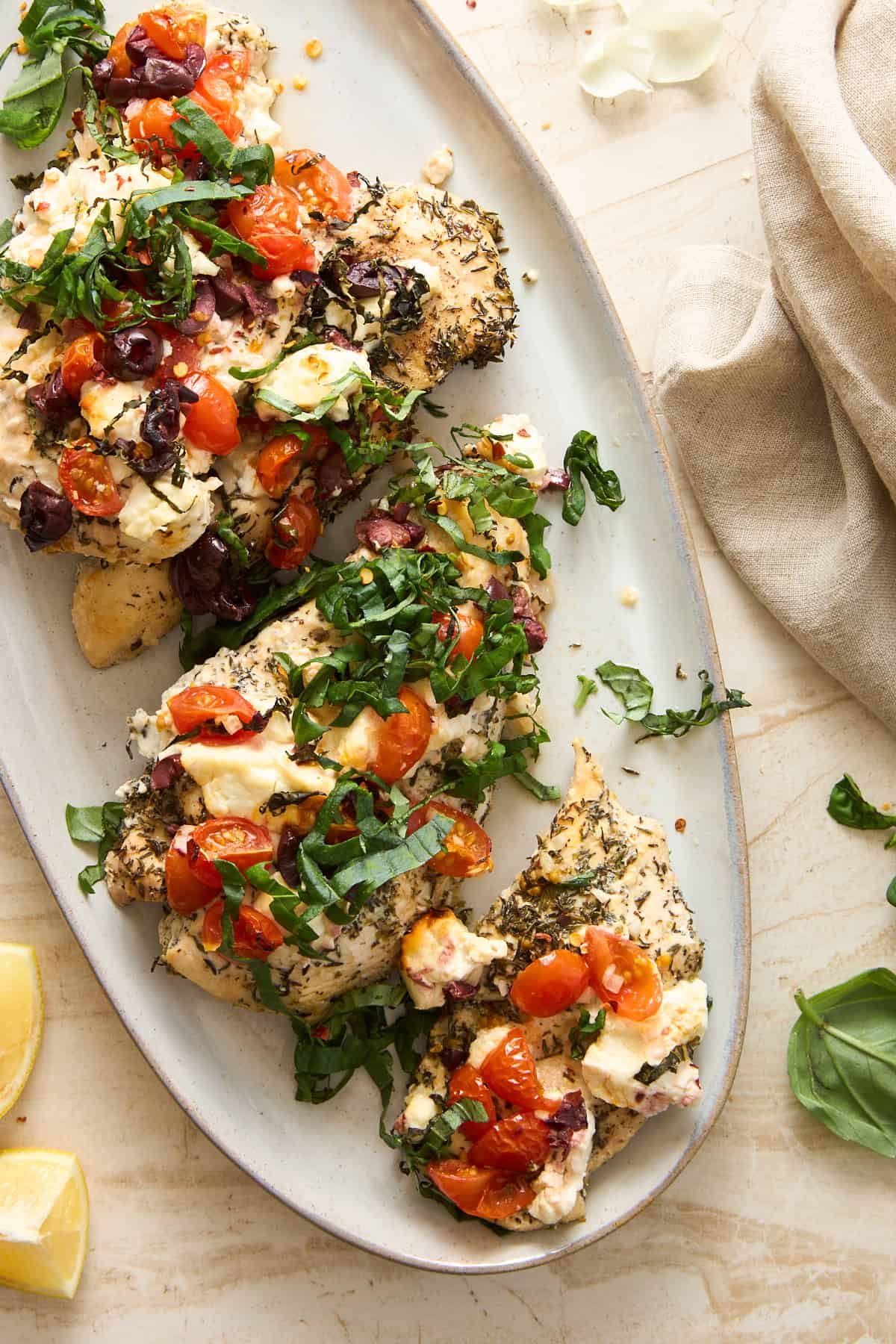 Herb-seasoned baked chicken breasts topped with melted cheese, cherry tomatoes, olives, and fresh chopped basil, served on an oval platter. Lemon wedges and basil leaves are nearby on a light-colored table.