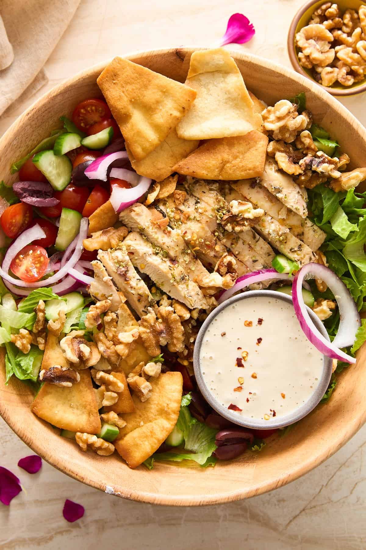 A wooden bowl filled with salad greens, grilled chicken slices, cherry tomatoes, cucumber, red onion, kalamata olives, walnuts, pita chips, and a small dish of dressing.