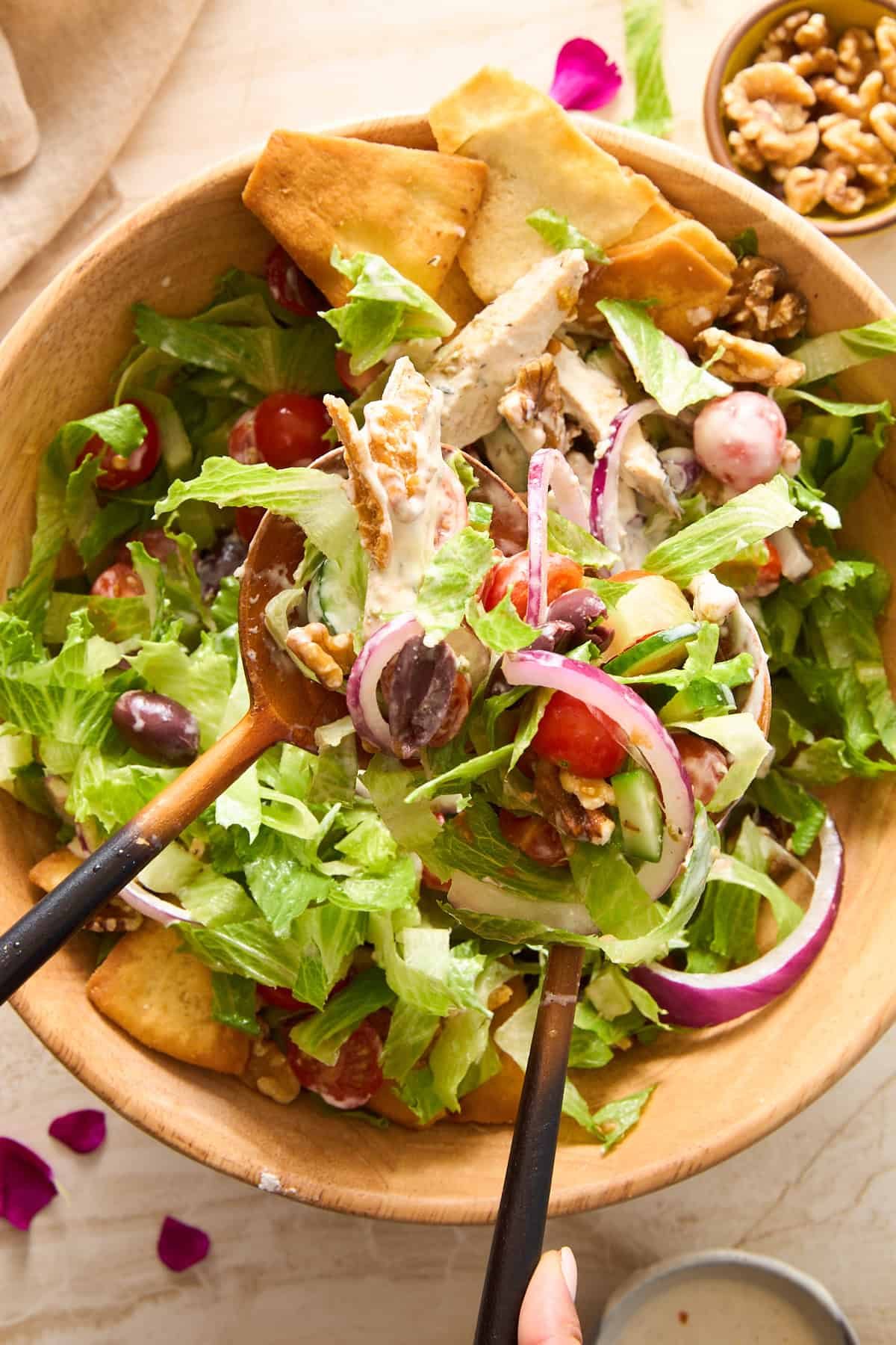 A wooden bowl filled with salad containing lettuce, grape tomatoes, red onion, olives, walnuts, pita chips, and creamy dressing. Two wooden serving spoons are tossing the salad. A small bowl of walnuts is nearby.