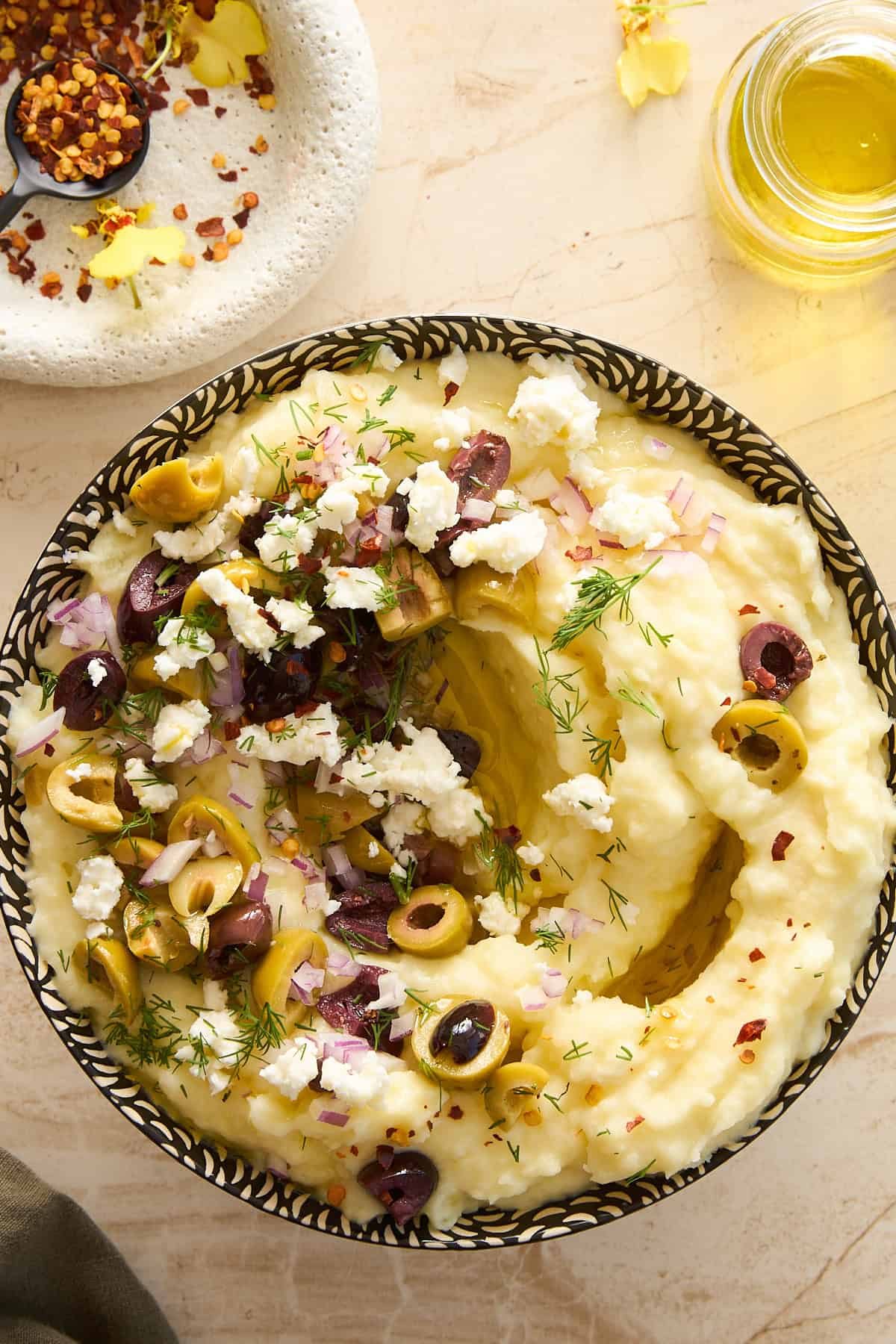 A bowl of mashed potatoes topped with chopped olives, crumbled feta cheese, red onions, dill, and a drizzle of olive oil, set on a light countertop next to a small plate of spices and a jar of oil.