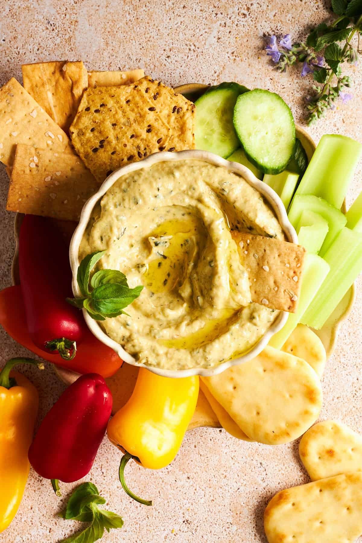 A bowl of zucchini dip on a platter with cucumbers, celery, pita, mini bell peppers, and crackers.