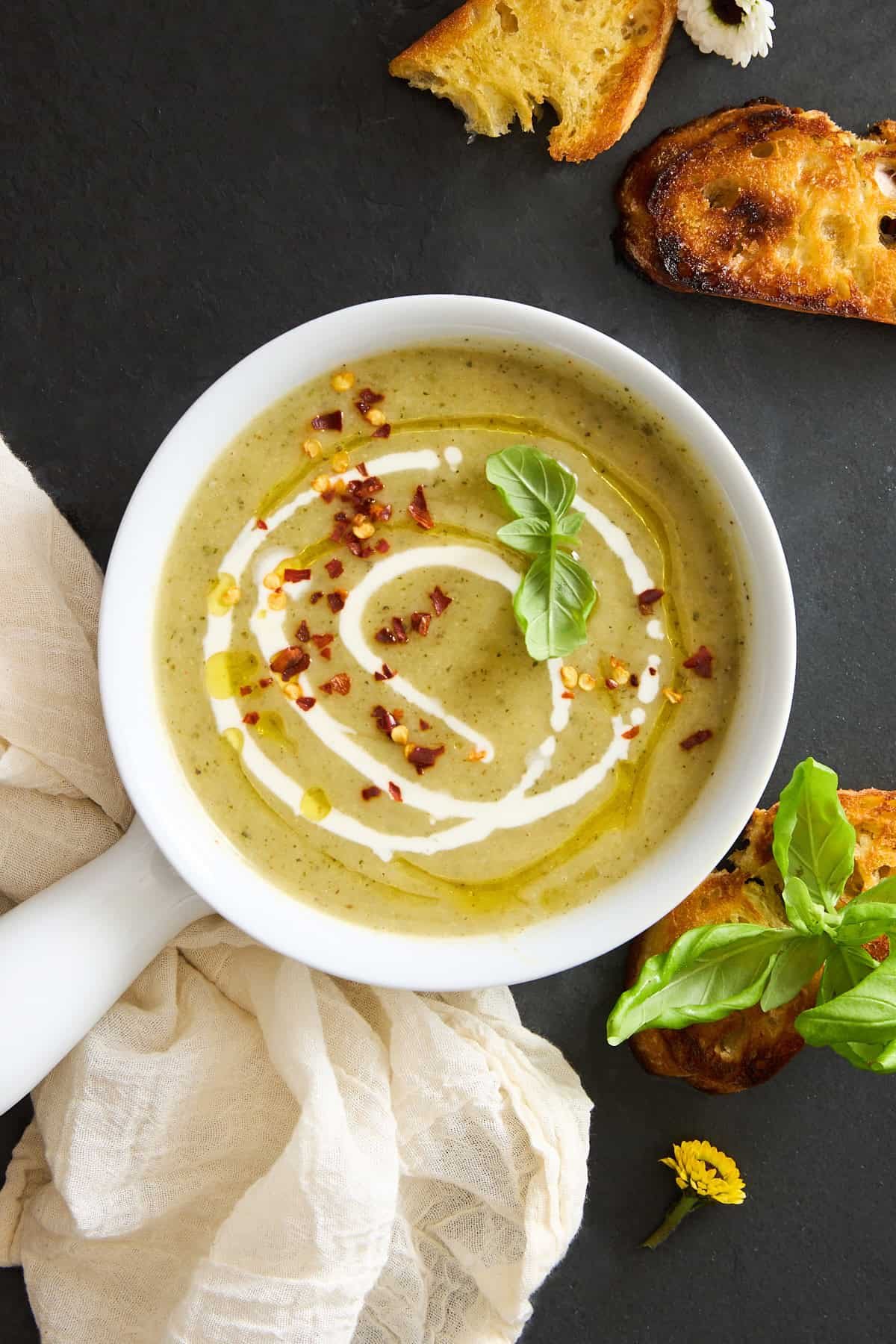 A bowl of silky green soup garnished with a swirl of cream, chili flakes, olive oil, and fresh basil, served with toasted bread slices on a dark surface beside a light cloth and a yellow flower.