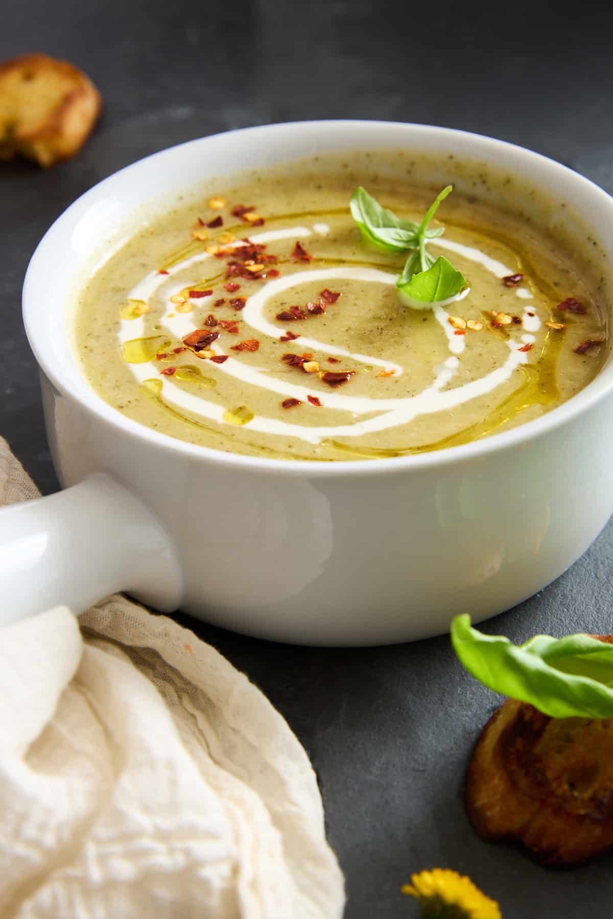 A bowl of creamy soup garnished with a swirl of cream, red pepper flakes, a basil leaf, and a drizzle of oil, served in a white bowl with a handle on a dark surface.