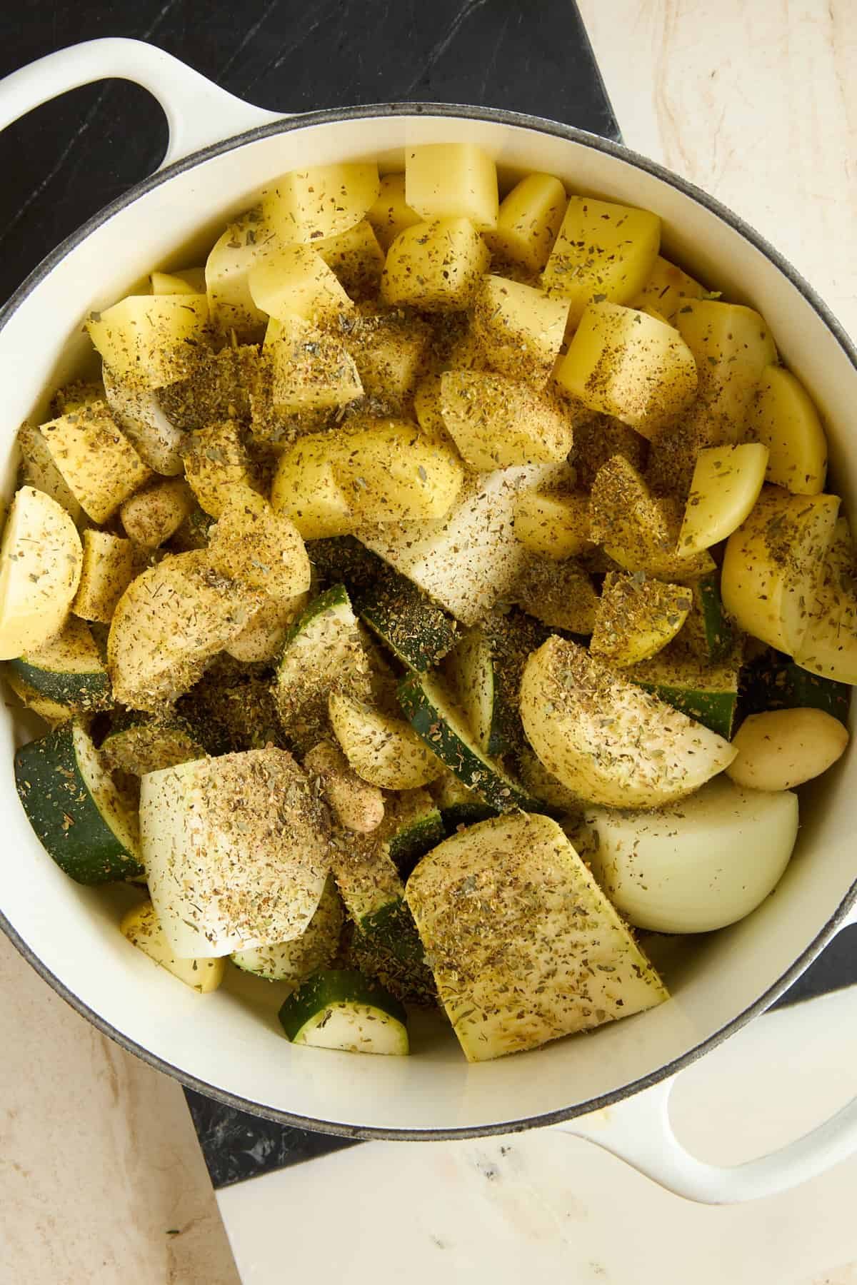 A white pot filled with chopped potatoes, zucchini, onions, and garlic cloves, all seasoned with a mix of dried herbs and black pepper, sits on a marble countertop.