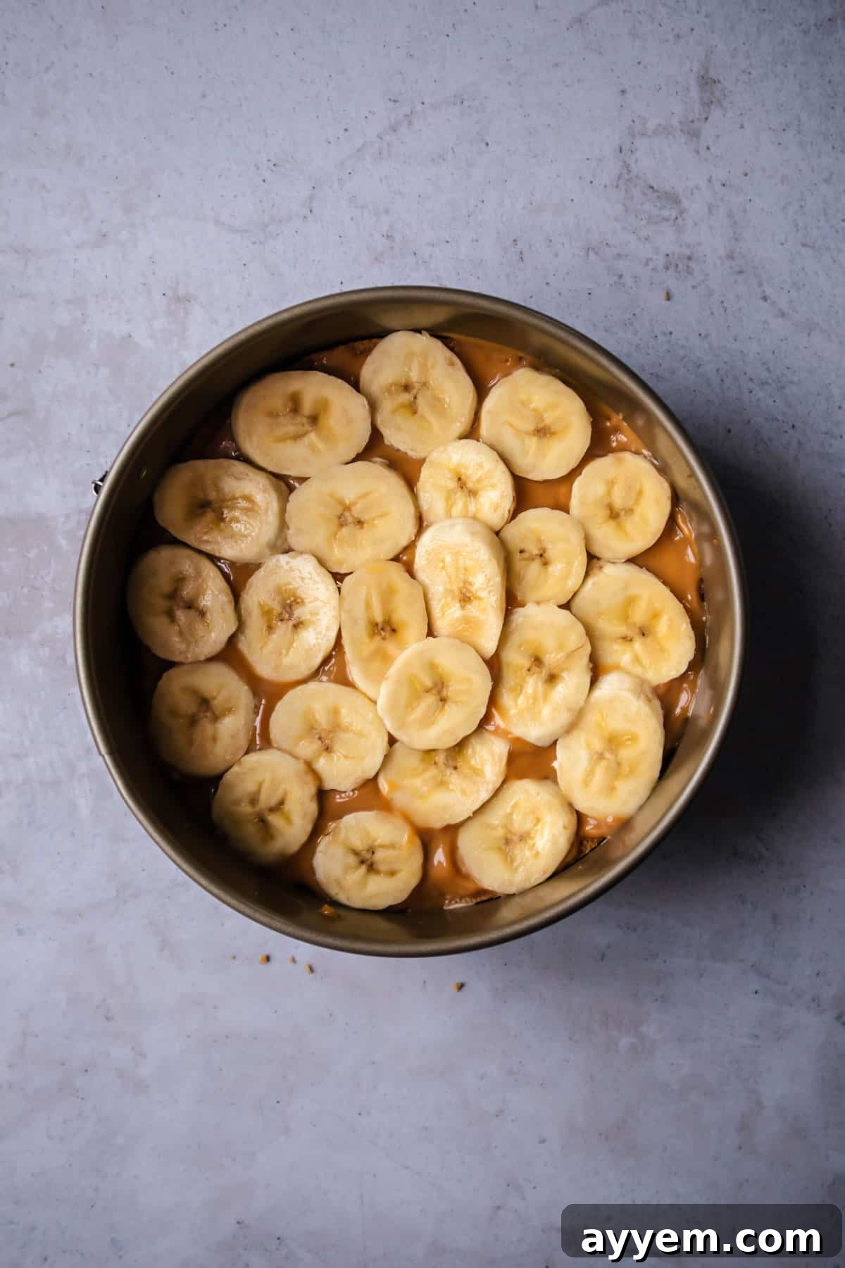 An overhead view of a cake pan featuring a beautiful graham cracker crust, a rich layer of dulce de leche, and perfectly arranged slices of fresh bananas, awaiting the creamy topping.