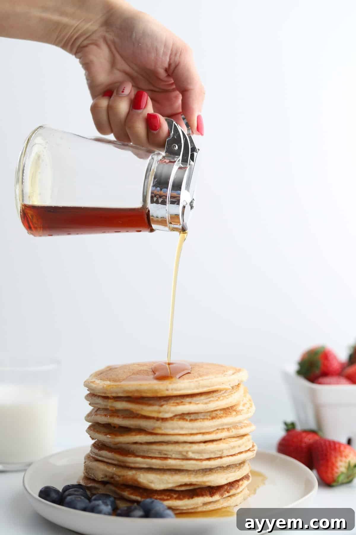 A hand pouring golden syrup over a tall stack of fluffy cottage cheese protein pancakes, garnished with fresh blueberries and a few extra berries scattered on the side, creating an inviting breakfast scene.