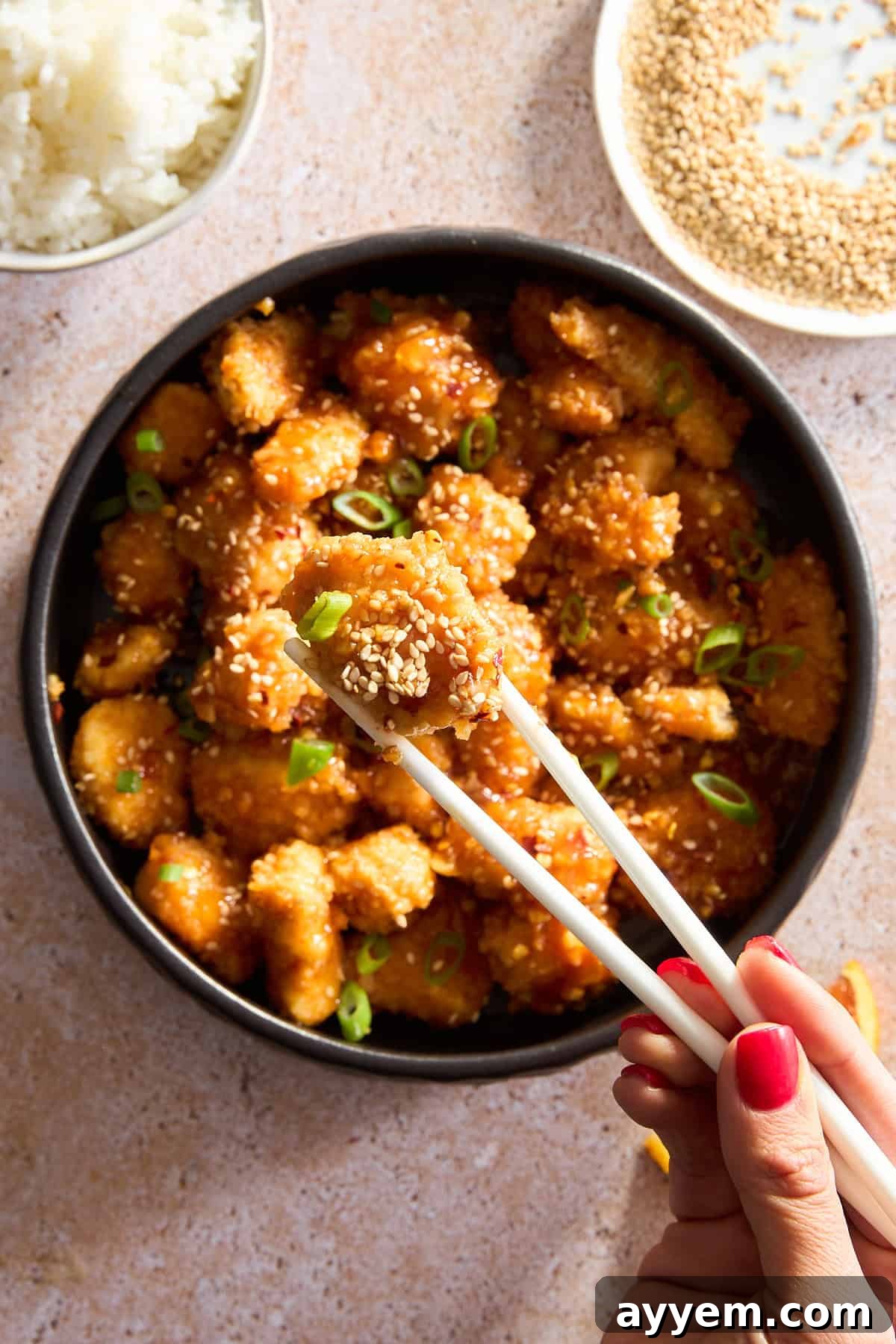Two chopsticks holding a piece of orange chicken over a bowl, garnished with sesame seeds and green onions.
