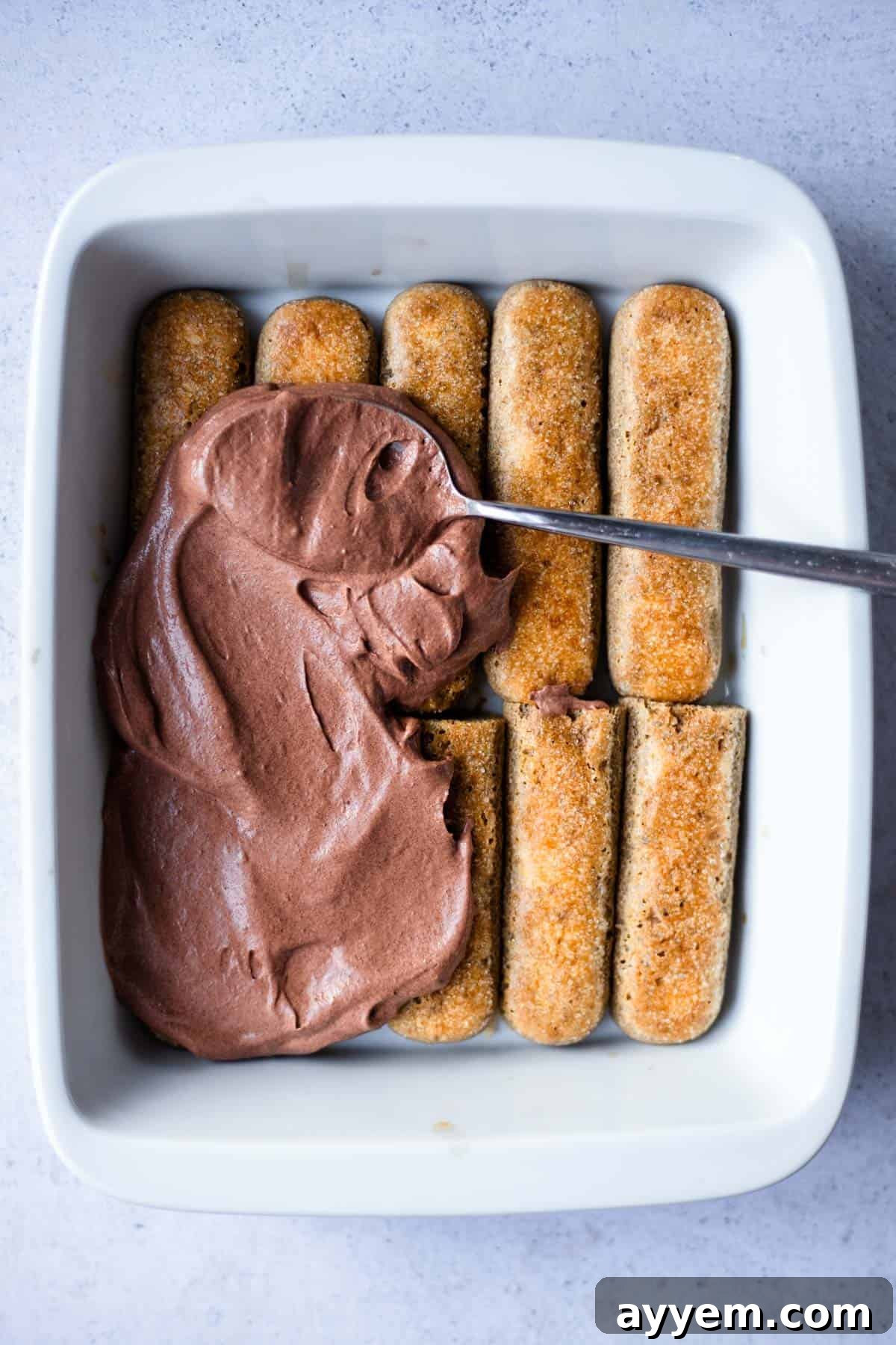 A smooth, rich layer of chocolate mascarpone cream being spread over the coffee-soaked ladyfingers in a baking dish.